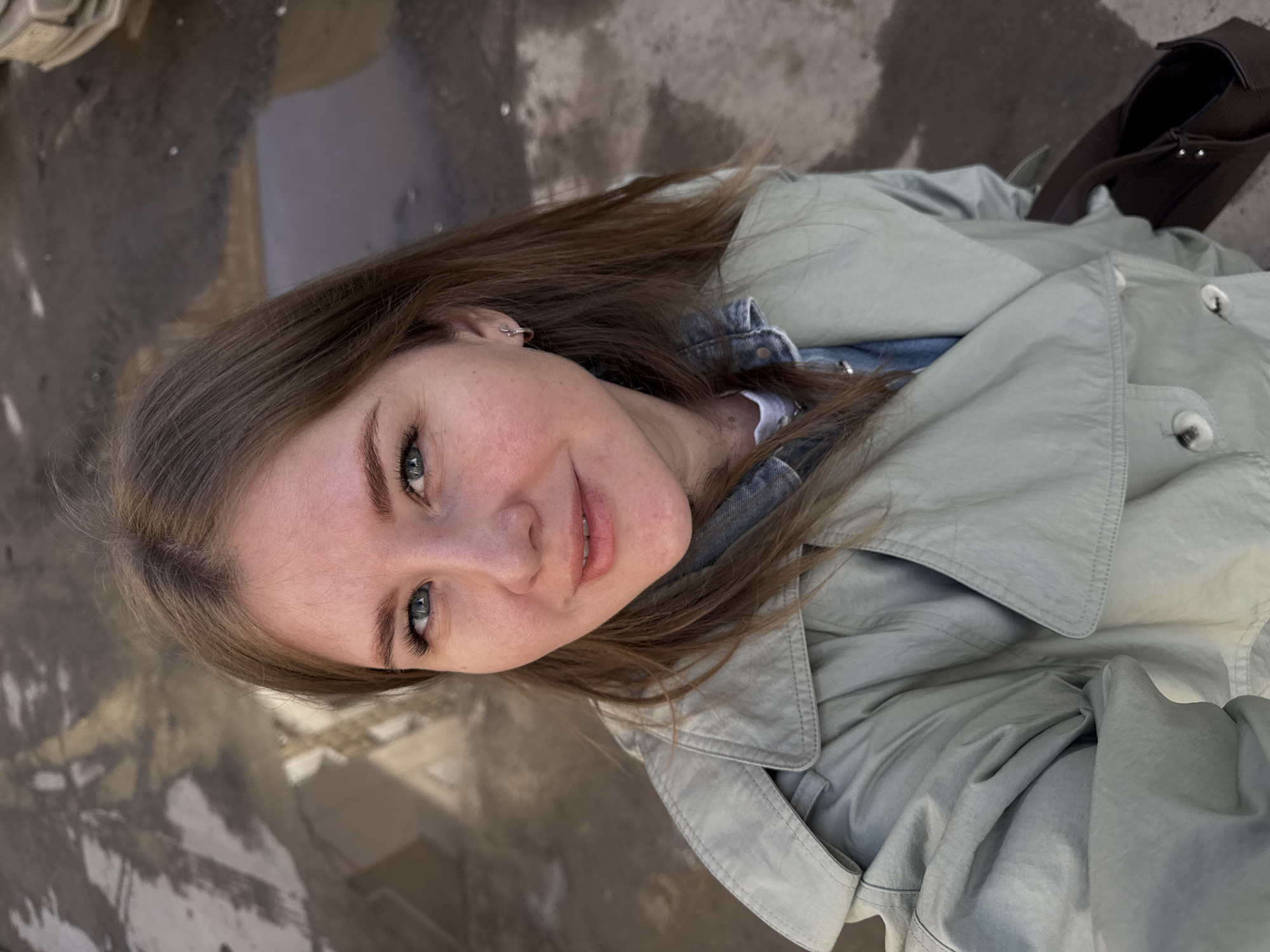 The image shows a young woman with long brown hair and blue eyes, wearing a light green jacket and a denim shirt, looking at the camera. The background appears to be a wet pavement, possibly reflecting the sky. The scene suggests an urban environment, consistent with the location of Perm, Russia. A dark brown bag is partially visible in the upper right corner of the image.