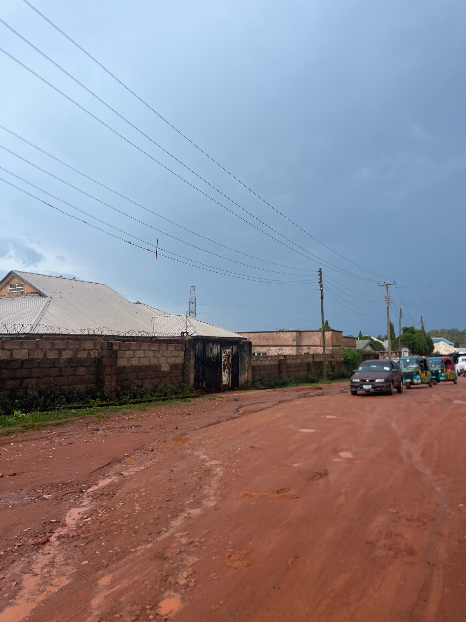 The image depicts a street scene in Jos, Nigeria, under an overcast sky with dark clouds suggesting impending rain. The road is unpaved, made of reddish-brown dirt, and shows signs of wear with numerous potholes and puddles of muddy water. Power lines are strung across the sky, supported by wooden and metal poles.
On the left side of the road, a brick wall topped with barbed wire encloses a property with a corrugated metal roof and a large, rustic metal gate. Behind the wall, a multi-pitched roof structure, possibly a residential building or commercial establishment, is visible. A high-voltage transmission tower stands in the background.
The right side of the road is occupied by a line of vehicles, including a dark-colored sedan and several auto-rickshaws, also known as Keke Napep, which are a common form of public transportation in Nigeria. The vehicles appear to be moving or stopped in traffic. Greenery, including grass and some shrubs, is present along the edge of the wall and the roadside. The overall impression is a typical urban or peri-urban environment in Jos, experiencing inclement weather.