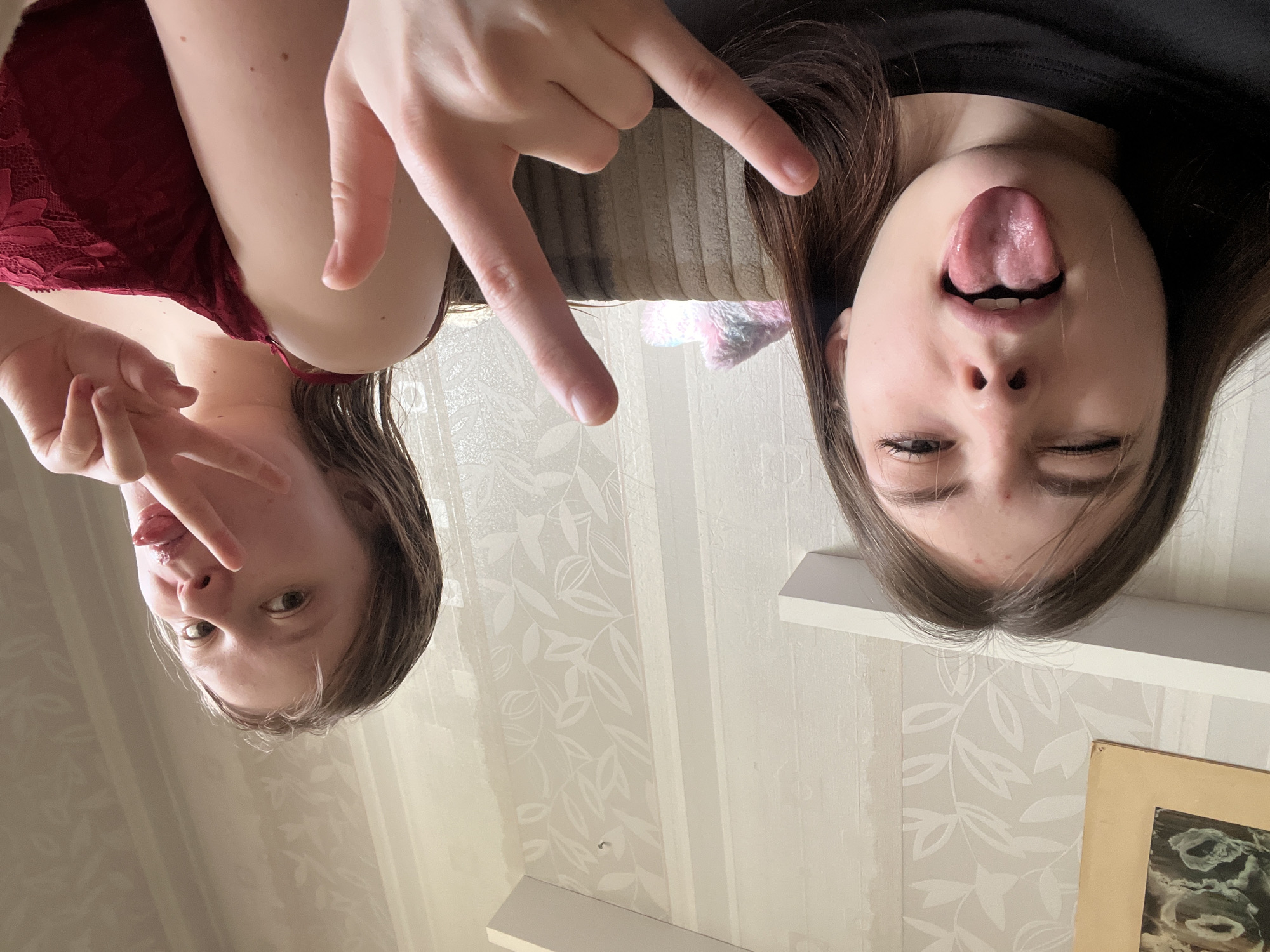 Two young women are posing for a selfie, lying on their backs, likely on a couch. The woman on the left is wearing a red top and making a peace sign with her fingers. The woman on the right is wearing a black top and sticking her tongue out, closing one eye. The location appears to be indoors, with patterned wallpaper and a picture frame visible. The city of Unknown, Russia is specified, although there is no contextual information in the image to confirm the location.