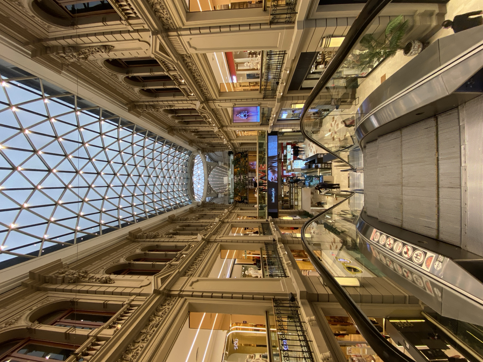 The image captures the interior of Galerías Pacífico, a shopping mall in Buenos Aires, Argentina. The camera is positioned looking up, emphasizing the architecture. A large, ornate ceiling with a geometric glass pattern allows natural light to flood the space. Multiple levels of retail spaces line both sides of the mall, with escalators providing access between floors. People can be seen on different levels, browsing or walking. The architectural details are elaborate, featuring decorative moldings, arches, and balconies. The escalator, with its glass railings and warning signs, is a prominent feature in the foreground.