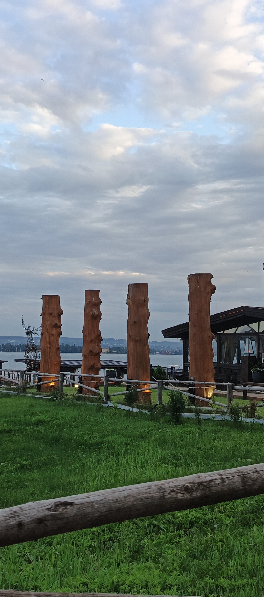 The image depicts a scenic outdoor view in Votkinsk, Russia, likely at dusk or during overcast conditions. The foreground features a lush green lawn with a rustic wooden fence. Behind the fence, four large, rough-hewn wooden pillars stand prominently, each illuminated at its base with warm-toned lights. To the left of the pillars, a slender, metallic sculpture, possibly an abstract human figure, is visible.
In the middle ground, a body of water, likely a lake or river, stretches across the scene. On the far side of the water, a distant shoreline with buildings and trees is discernible. Adjacent to the wooden pillars and partially obscuring the water is a modern structure with a dark roof and glass enclosures, suggesting a restaurant or cafe. The sky above is filled with a mix of light and dark clouds, with hints of blue sky peeking through. The overall atmosphere is serene and tranquil, with the lighting on the pillars adding a touch of warmth to the cool-toned environment.