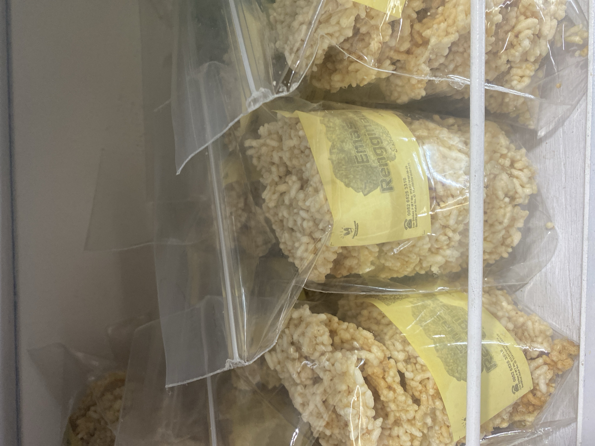 The image shows several packages of a crunchy, puffed rice snack, likely a type of rice cracker, stacked in what appears to be a retail display or storage area, possibly a freezer or refrigerator unit due to the white vertical support bar visible. The snacks are translucent through their clear plastic packaging, revealing their irregular, porous texture and light brown, golden color.
Each package has a bright yellow label with text printed in dark lettering. The visible text on the labels includes "Emas Renggina," "Since 198," and a phone number "0852 6525 3318." There are also Indonesian regulatory markings like "Izin Dinkes PIRT No. 2151171010138-25" and "Sertifikal Halal No. ID 11410033010301125," indicating food safety and halal certification.
The lighting is somewhat dim, and the focus is on the products. The overall impression is a commercial setting where food items are being presented or stored. The image is taken from a slightly angled perspective, looking down and across the stacked packages. There are no people or animals visible.