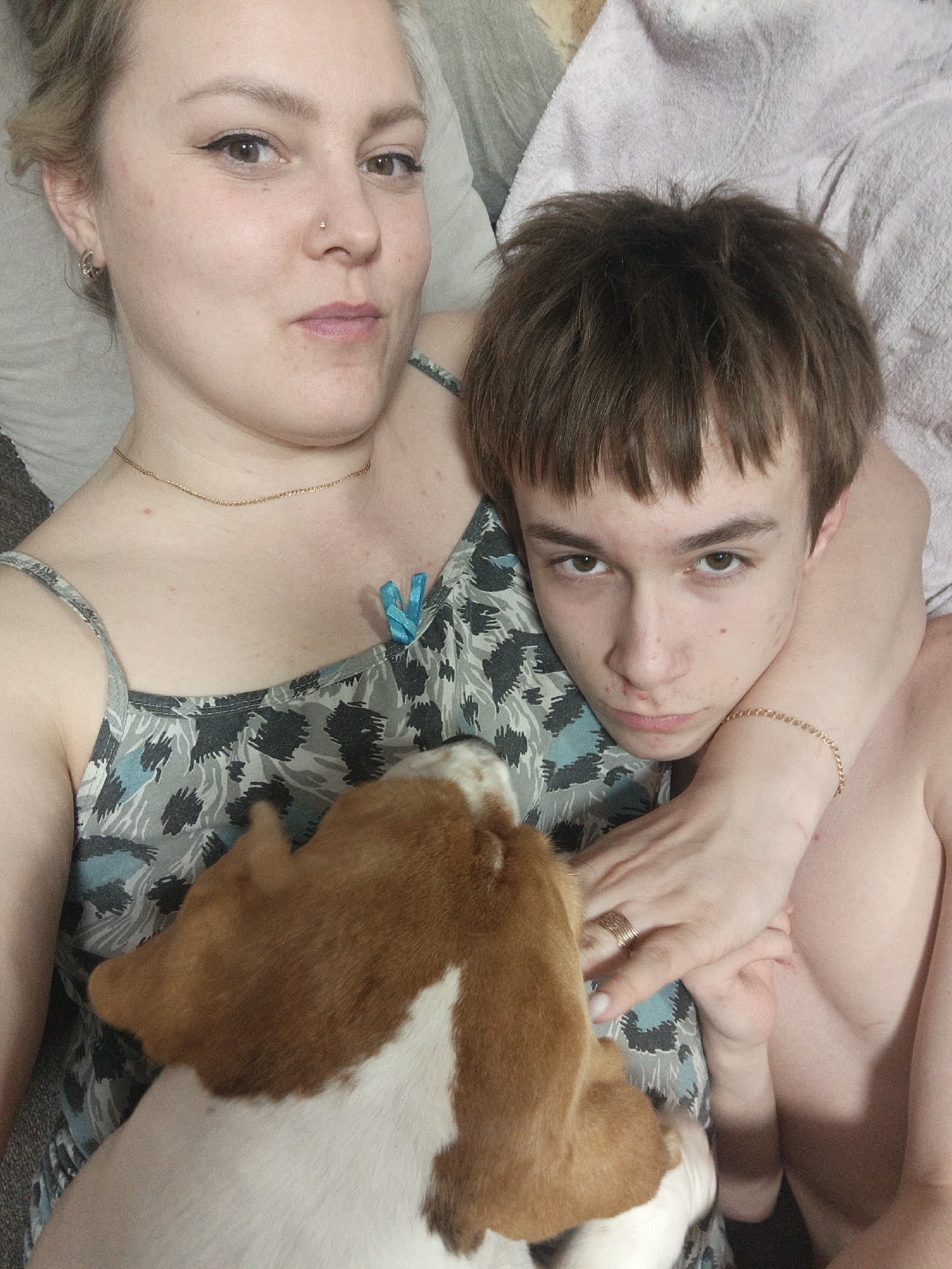 A photograph taken indoors depicts two people, a dog, and surrounding textiles. The adult female on the left, wearing a patterned tank top and a gold necklace, has a fair complexion, light brown hair pulled back from her face, and a nose piercing. Her expression is neutral. A younger male lies behind her, with his bare arm draped across her shoulders. He also has fair skin, brown hair in a short, layered style, and a serious expression. A brown-and-white dog, possibly a beagle, is nestled between them, facing away from the camera. The background consists of soft textiles, likely a couch or bed, with a light pink blanket partially visible on the right side. The precise location in the city of Марковский, Russia, cannot be determined from the image.
