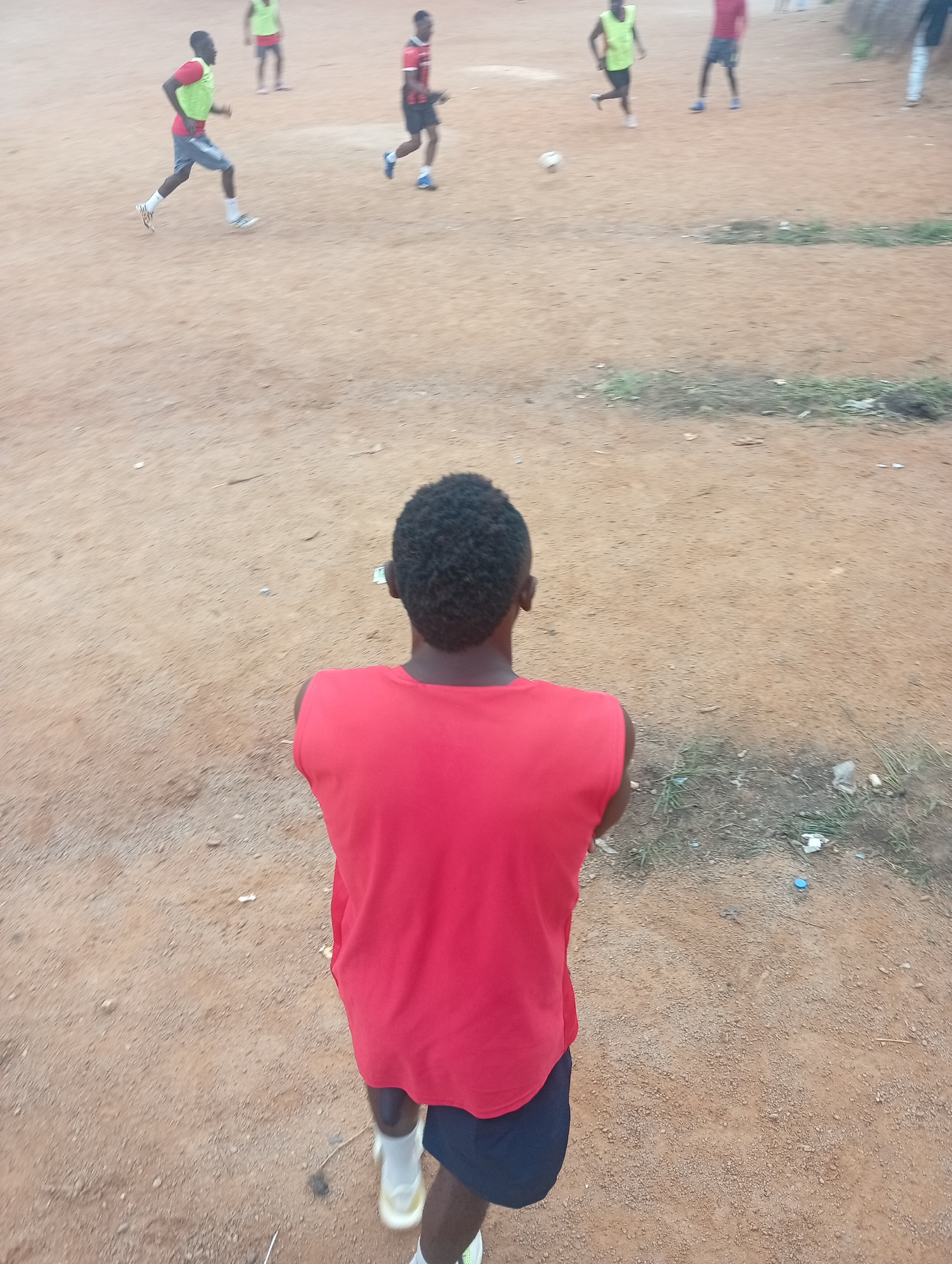 An over-the-shoulder view captures a young Black male from the chest down, wearing a sleeveless red shirt and dark blue shorts. He stands on a dirt field, possibly a football pitch, in Jos, Nigeria. His white socks and athletic shoes are visible. In the background, several other young Black males are engaged in a football game. One player, wearing a red shirt and lime green vest, is running with the ball. Another player in a red shirt and blue shorts is positioned nearby. The environment is a dry, dusty field with scattered patches of dry grass and some litter. The lighting suggests it is daytime, likely late afternoon or early evening given the soft light.