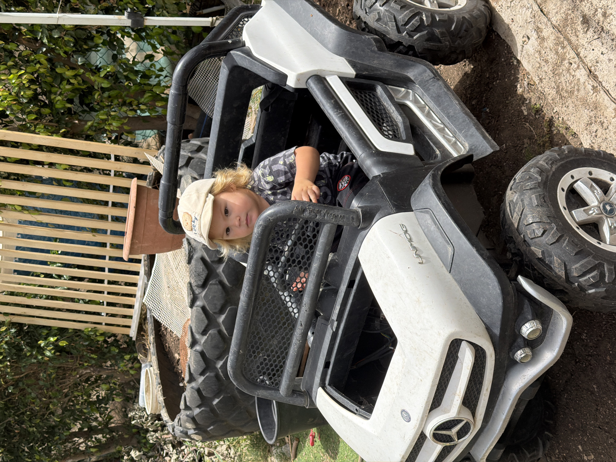 A young child wearing a baseball cap is sitting inside a white and black toy vehicle, potentially a miniature all-terrain vehicle (ATV) or utility terrain vehicle (UTV). The vehicle is positioned outdoors on a surface of dirt and possibly grass. The background features a fence, greenery, and other outdoor elements. The exact location in Kiryat Bialik, Israel, cannot be determined from the image.