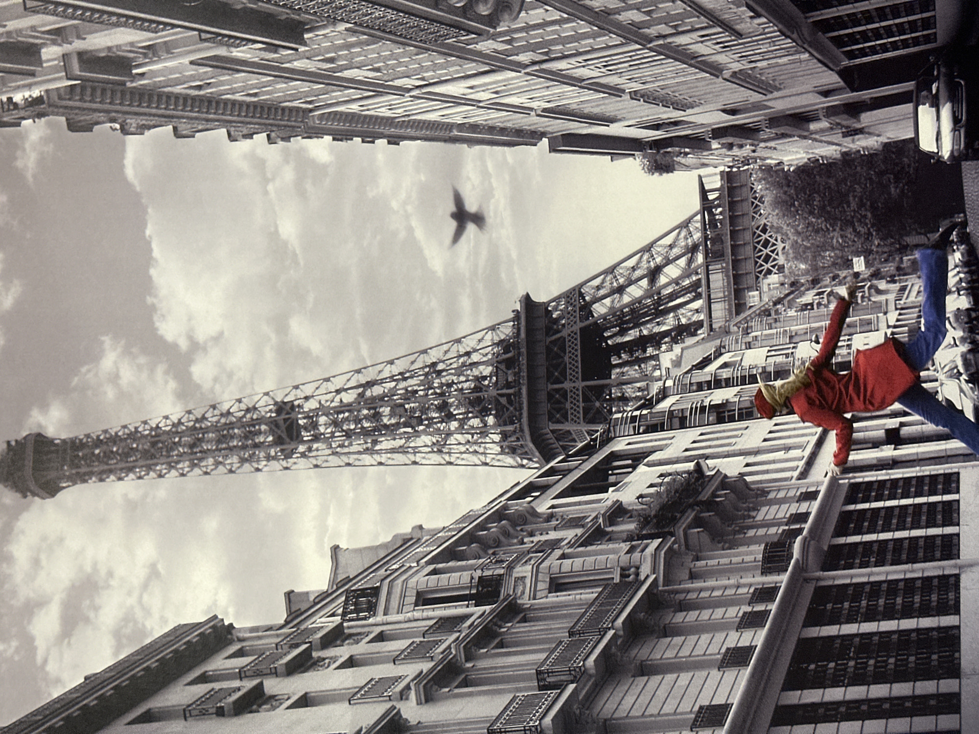 The image depicts a black and white scene with a single subject, a woman in a red coat and blue pants, jumping in mid-air. She is positioned in the lower left quadrant of the frame, with her arms extended. Behind her, to the left, is the Eiffel Tower, a prominent landmark. The buildings flanking the street are typical Parisian architecture, with intricate details and balconies. In the sky, a bird is captured in flight. The street below shows parked cars. The location is not Energetik, Russia, but rather Paris, France.
