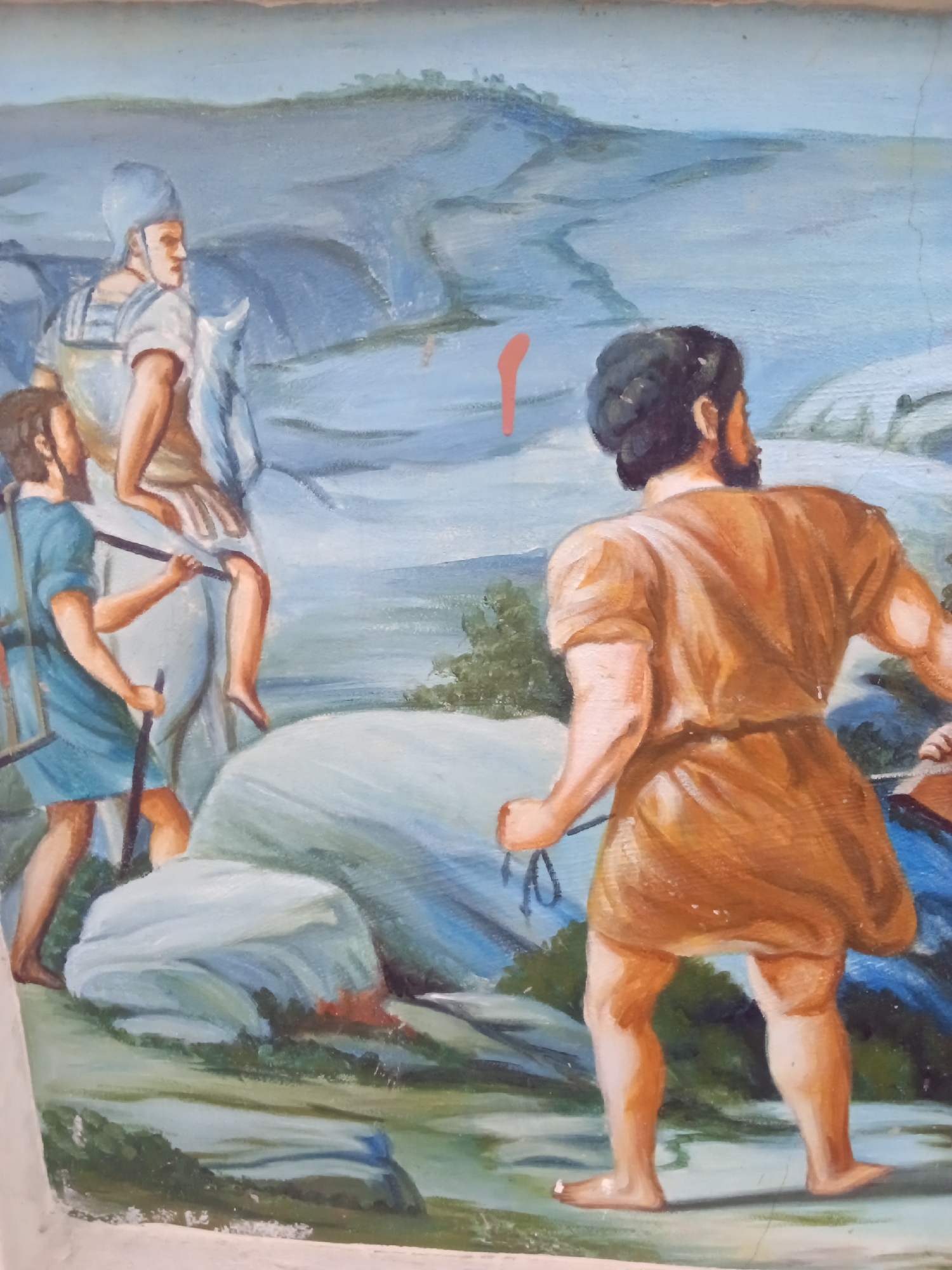 The painting depicts a scene involving multiple figures in what appears to be a historical or biblical context. In the foreground, a muscular man with dark hair and a beard stands with his back to the viewer. He is wearing a brown tunic and holds a sling in his hand. He appears to be facing a group of soldiers. To the left, there are two figures, one mounted on a light-colored horse and the other walking alongside. The mounted figure wears armor and a helmet. The figure walking beside the horse wears a blue tunic, carries a shield and spear, and appears to be looking up at the mounted soldier. The background features a landscape with hills and a body of water. The painting's overall style is reminiscent of historical or religious illustrations. The scene is depicted with visible brushstrokes. The location is Ado, Nigeria.