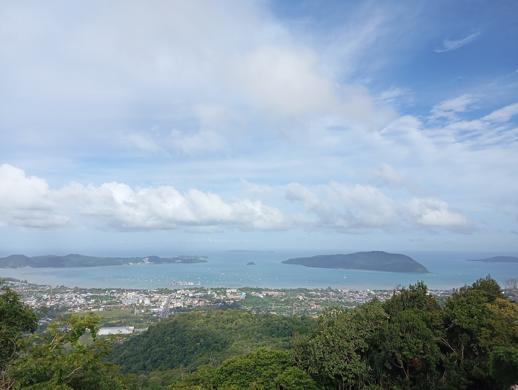 This is a high-angle, wide-shot photograph taken from a viewpoint above Karon, Thailand. The image captures a panoramic view of the city, the bay, and the surrounding islands under a partly cloudy sky. In the foreground, dense green foliage, including trees and shrubs, frames the lower portion of the scene. Below the foliage, the city of Karon spreads out, characterized by numerous buildings, some with white and red roofs, and a road system visible. The city meets a wide, calm bay filled with a multitude of sailboats and yachts anchored or moving slowly. The bay extends into the distance, where several islands of varying sizes and elevations are visible. The largest island in the middle ground has a distinct, rounded shape. The sky is a pale blue with scattered white cumulus clouds, some of which are thin and wispy, while others are larger and more defined. The overall atmosphere is bright and serene, suggesting a clear day with moderate cloud cover.