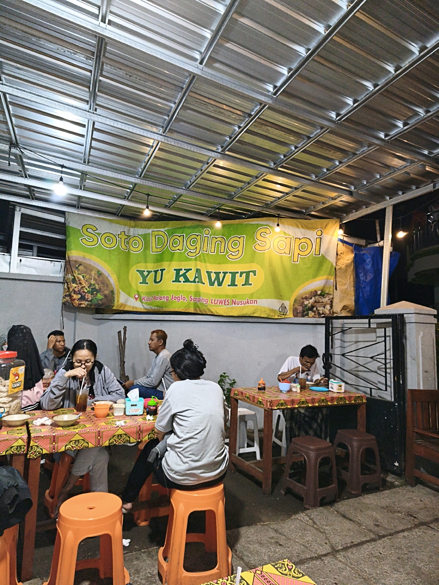 The image depicts an outdoor dining area of a restaurant in Surakarta, Indonesia, with a banner that reads "Soto Daging Sapi YU KAWIT Kidul Palang Joglo, Samping LUWES Nusukan". Several people are seated at tables, eating and interacting. In the foreground, a woman in a gray jacket is drinking from a glass with a straw, while another person in a gray shirt sits behind her. To the right, a man in a white shirt is eating at a table. The setting appears to be casual, with simple tables and plastic stools. The roof is made of corrugated metal, supported by a metal frame. The overall atmosphere suggests a local eatery.
