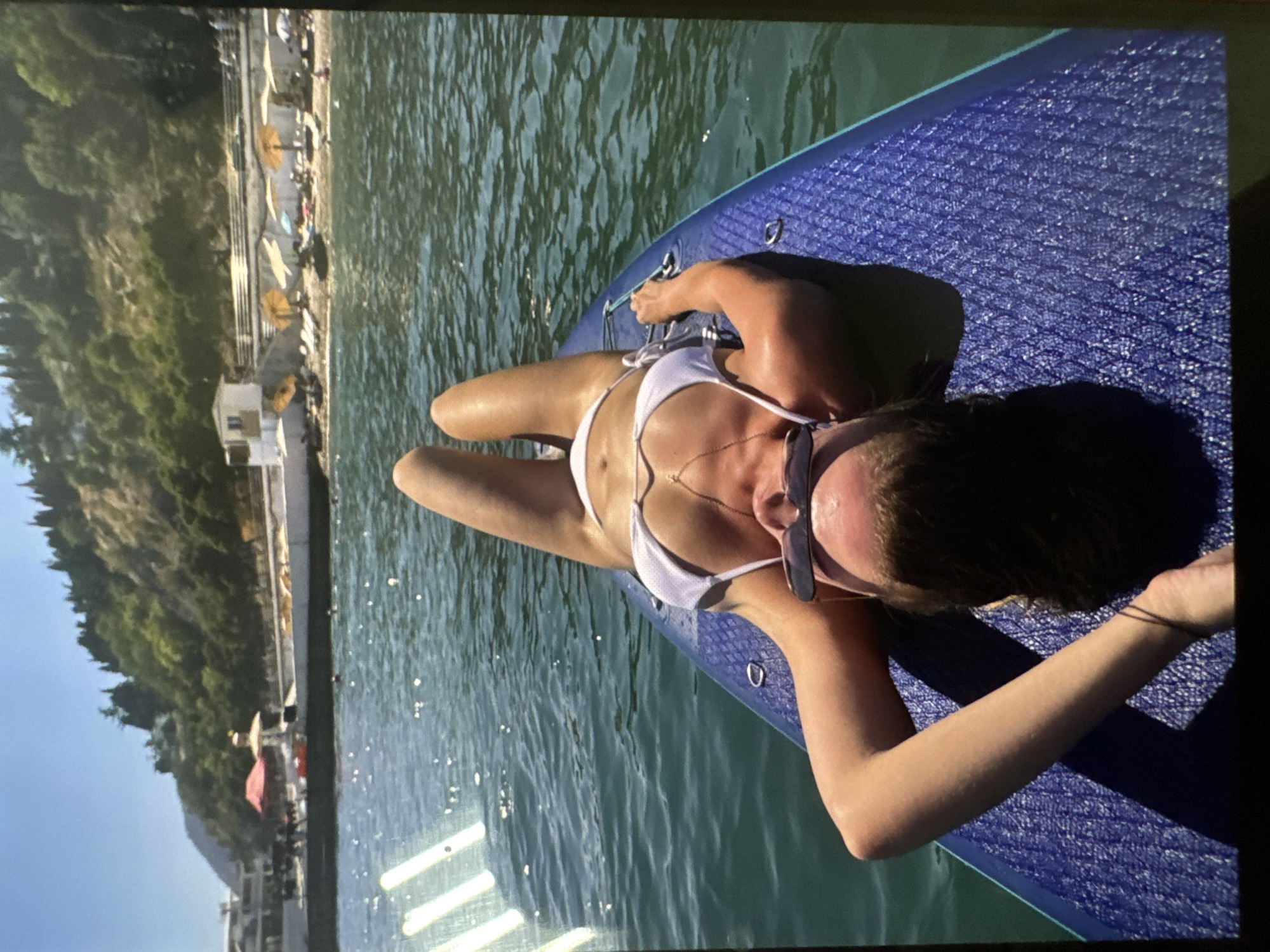 A woman in a white bikini lies on her back on a blue paddleboard in the ocean. Her legs are raised, and her arms are resting behind her head. She is wearing sunglasses. In the background, a beach with umbrellas and lounge chairs is visible, backed by a tree-covered hill. The water is calm and blue, and the sky is clear.