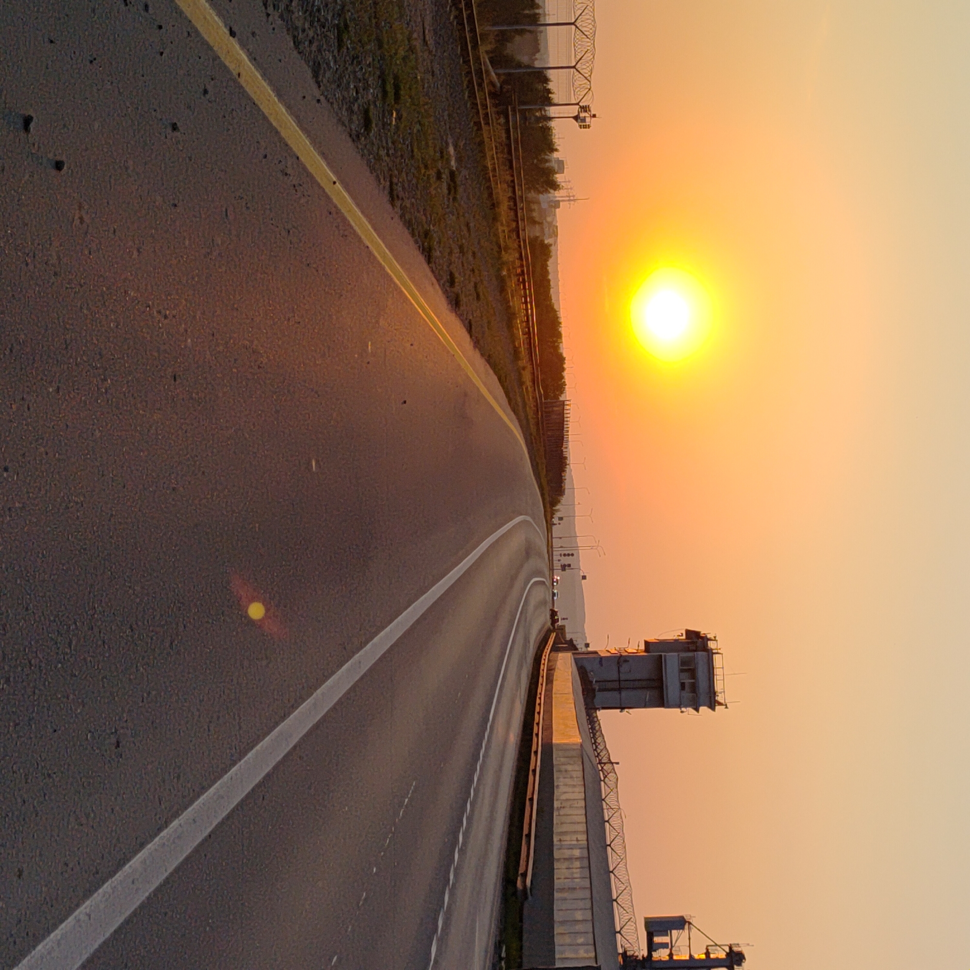 The image depicts a wide asphalt road curving to the right under a hazy orange sky during sunset. A bright, diffused sun is visible in the upper left quadrant. The right side of the road features a modern concrete barrier with barbed wire on top, and a tall, multi-story control tower structure. The left side of the road has a gravel shoulder with some sparse vegetation and railroad tracks running parallel. In the distance, along the horizon, a few more industrial structures and what appear to be wind turbines are faintly visible. There are no people or vehicles clearly discernible in the foreground or mid-ground. The overall scene suggests an industrial or transportation hub on the outskirts of Chaikovsky, Russia, at dusk.