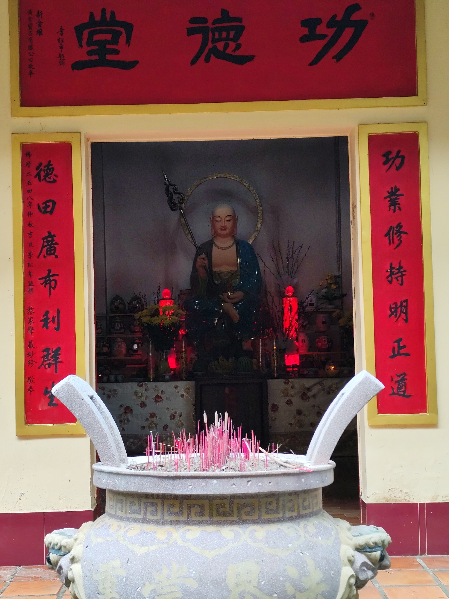 A close-up, medium shot depicts a statue of a Buddha figure within a shrine, with a large incense burner in the foreground, in Da Lat, Vietnam. The Buddha statue, positioned centrally within the shrine, is seated and holds a tall staff. The backdrop of the shrine is adorned with various religious artifacts and floral arrangements, illuminated by red lights. A large, ornate gray stone incense burner dominates the foreground, filled with numerous lit pink incense sticks. The shrine is framed by pillars displaying red banners with traditional Chinese calligraphy. Above the shrine is a red banner with larger Chinese characters. The structure itself is painted in a pale yellow color, with a red base. The scene is brightly lit, emphasizing the Buddha statue and the burning incense sticks.