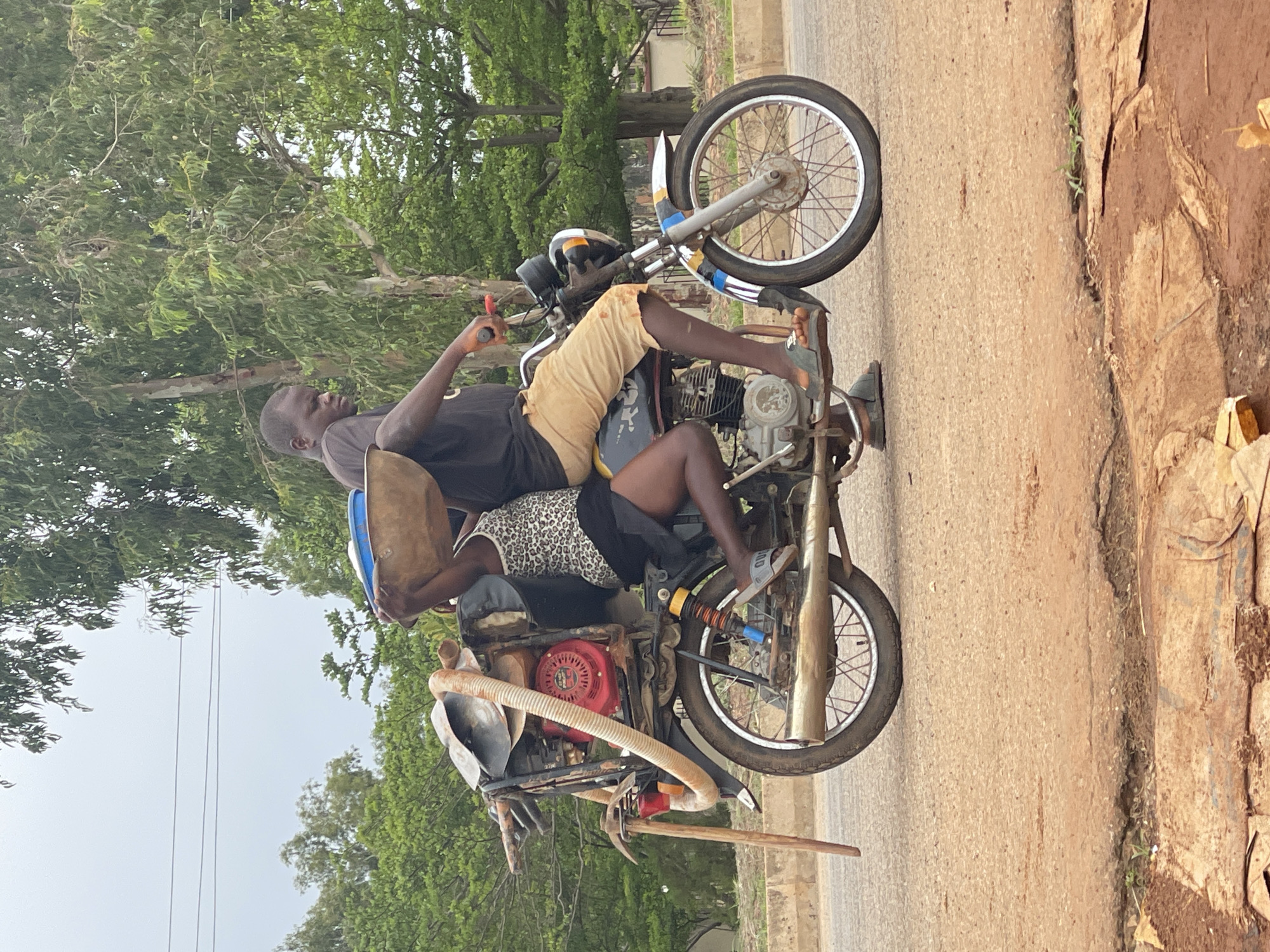 The image shows two individuals on a motorcycle. The person in the front appears to be a male, wearing a dark t-shirt and light-colored shorts. He is operating the motorcycle. The person behind him, possibly a female, is wearing a patterned tank top and dark shorts. She is holding a large, shallow basket with a blue lid on top. The motorcycle is loaded with various items on the rear, including what appears to be a red engine component, a metal rack, and a long wooden object resembling a tool, possibly a hoe. The motorcycle is on a paved road, which shows signs of wear and tear, with cracks and patches. The surrounding environment is lush with trees and vegetation. The sky is overcast. The location is identified as Ban, Nigeria.