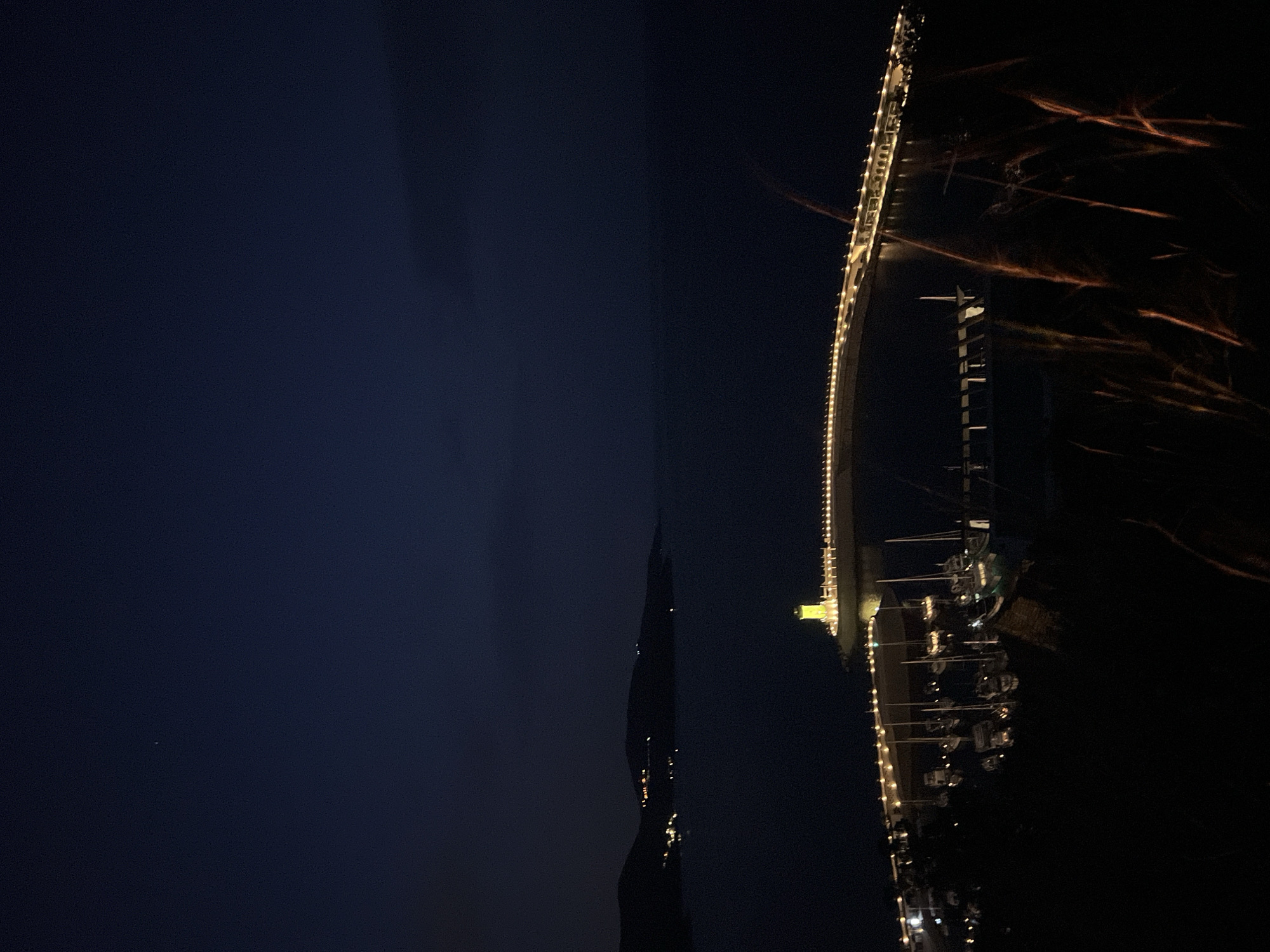 At night, the image captures the illuminated Porto Montenegro marina in Tivat, situated on the Bay of Kotor in Radovići, Montenegro. The marina is lined with yachts, lit by dockside lighting. The night sky is dark, and there are what appear to be light streaks above and beyond the marina.