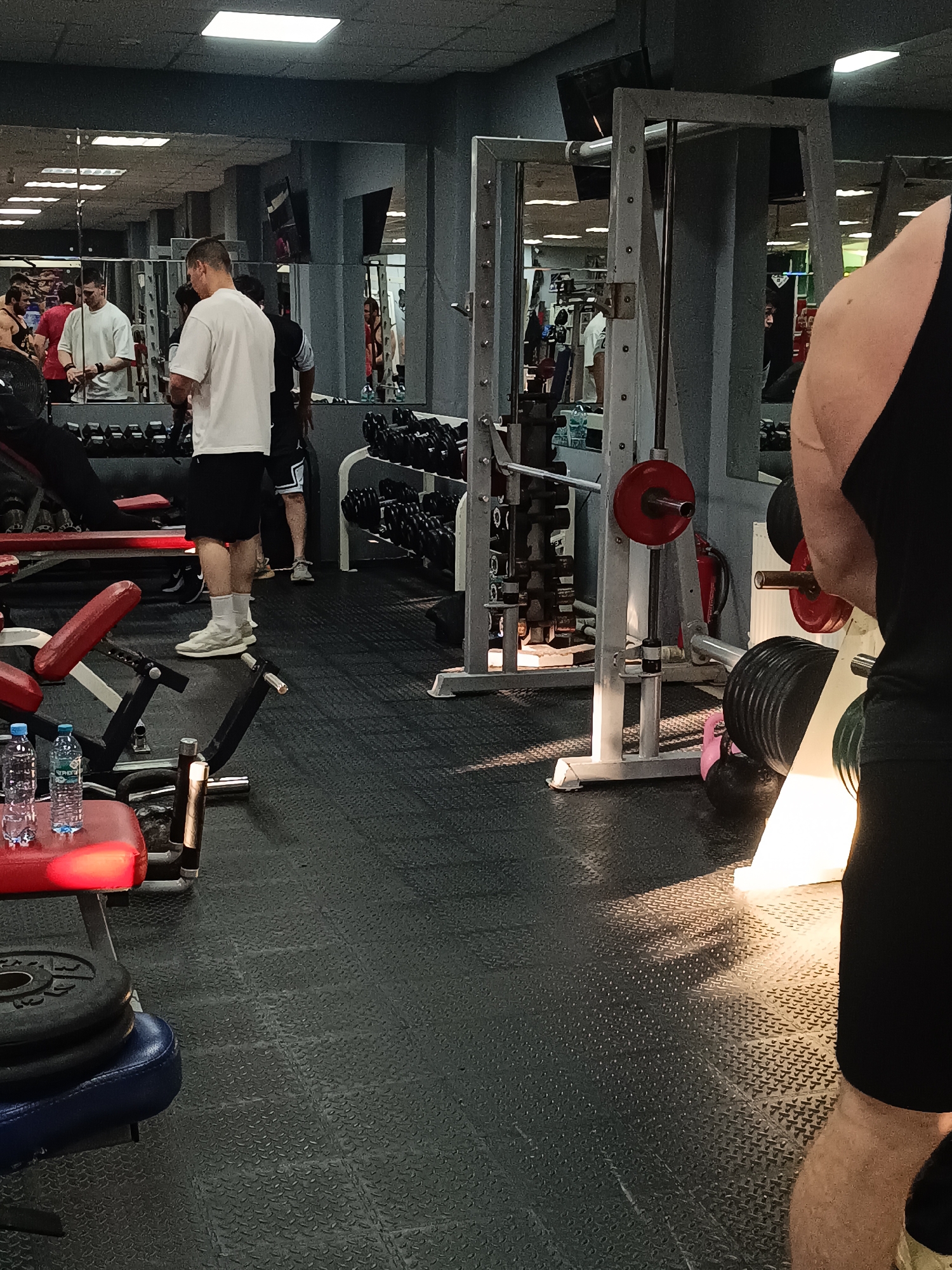 This image depicts an interior view of a bustling gym or fitness center in Naberezhnye Chelny, Russia. Several men are present, engaged in or preparing for weightlifting activities.
In the foreground on the right, the muscular right arm and torso of a man wearing a black tank top and black shorts are visible. A strong beam of natural or artificial light illuminates the textured black rubberized floor tiles near his legs.
Towards the center, a man wearing a white t-shirt, black shorts, white socks, and light-colored sneakers stands with his back partially to the camera, facing a wall of mirrors. His reflection, along with those of other gym patrons in various workout attire (some shirtless, others in t-shirts or tank tops), is visible in the mirrors, suggesting a busy environment.
The gym is well-equipped with various weightlifting apparatus. Multiple dumbbell racks laden with numerous dumbbells of different sizes line the walls. A prominent silver squat rack or Smith machine with a barbell and red weight plates is situated centrally. A red fire extinguisher is visible behind this equipment. On the left side, there are red and blue padded benches, with black weight plates stacked nearby. Two clear plastic water bottles with blue labels, one partially obscured, rest on a red bench in the lower-left corner. A pink and a black kettlebell are visible on the floor near the squat rack.
Overhead, square fluorescent panel lights illuminate the gym. Visible text includes numbers on the weight plates, such as "30" on a black plate and "25" on red and black plates, likely indicating weight in kilograms. The scene conveys an atmosphere of active physical training.