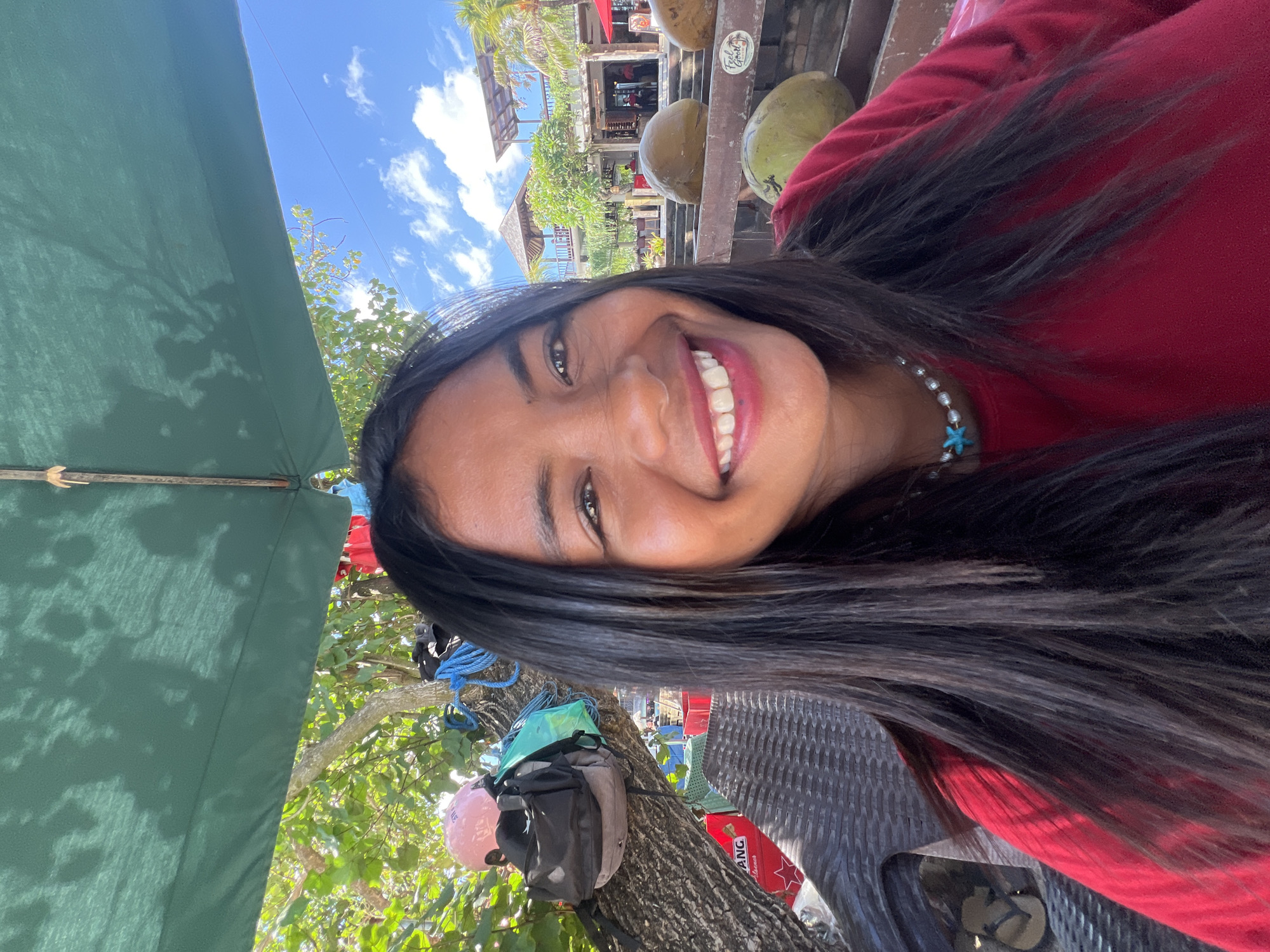 A woman with long, dark hair and a bright smile is pictured in a close-up selfie. She is wearing a red shirt and a pearl necklace with a blue starfish pendant. In the background, there is a tropical scene with lush greenery, palm trees, and buildings that appear to be part of a resort or restaurant. Several coconuts are visible in the foreground, resting on a wooden railing. The woman is seated under a large green umbrella, suggesting an outdoor setting, possibly a beachside cafe or bar in Kuta, Indonesia. The sky is a clear blue with scattered white clouds. The overall atmosphere is relaxed and sunny, characteristic of a vacation destination.
