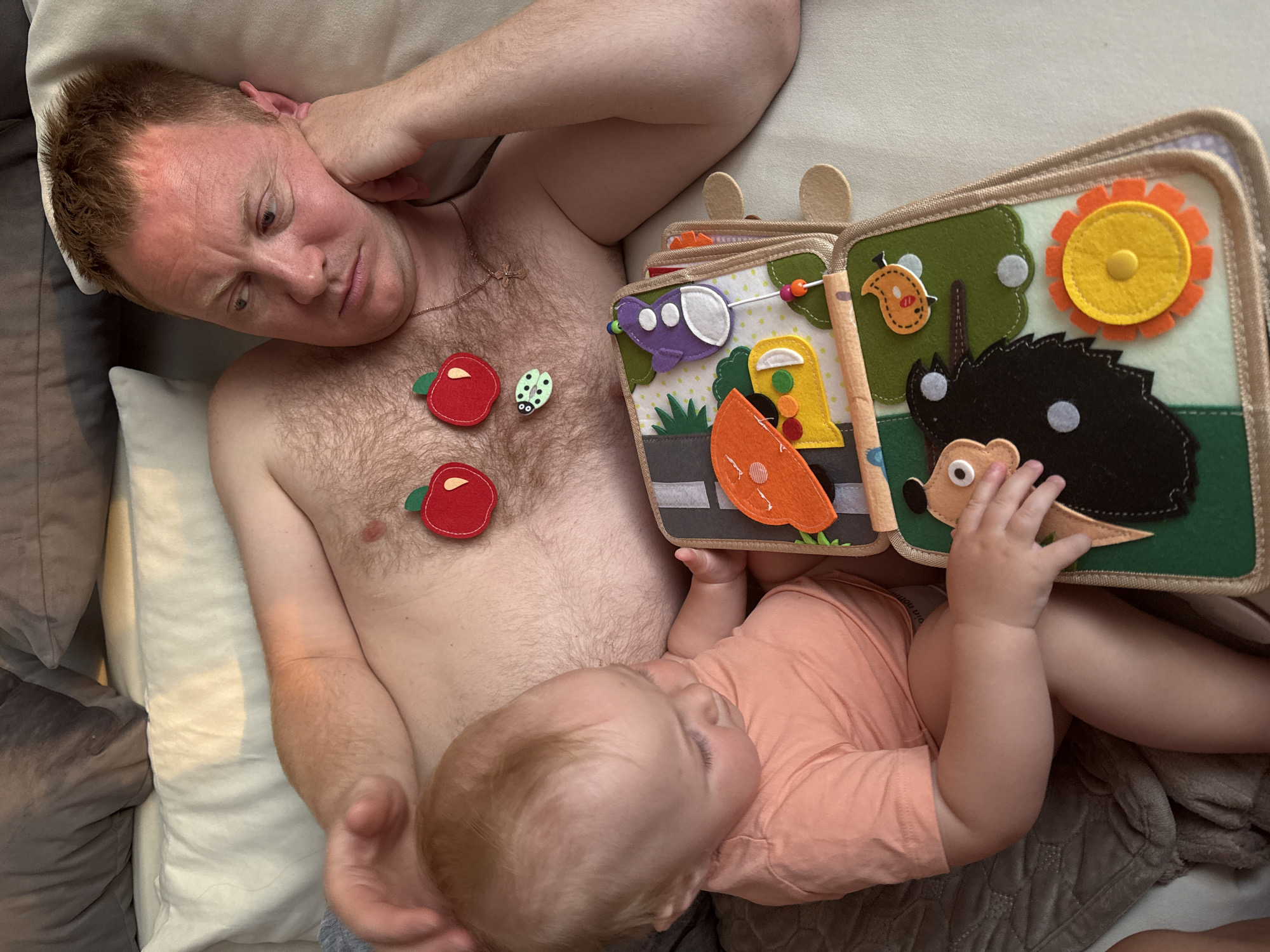 This overhead shot captures a man and a baby lying on a couch. The man, shirtless, has his hand behind his head and appears to be looking at the baby with a pensive expression. A few felt appliques, shaped like red apples and a ladybug, are attached to his chest. Next to him, a baby in a peach-colored shirt is leaning on the man, engrossed in a felt book. The book features various colorful felt elements, including a hedgehog, a sun, a bird, and a vehicle. The baby's hand is reaching out towards the book. The scene takes place indoors on a couch, likely in a home setting. The lighting suggests it could be daytime. The overall mood is intimate and peaceful, hinting at a moment of shared playtime or quiet connection between father and child. No visible text is present in the image. The location is Chaikovsky, Russia.