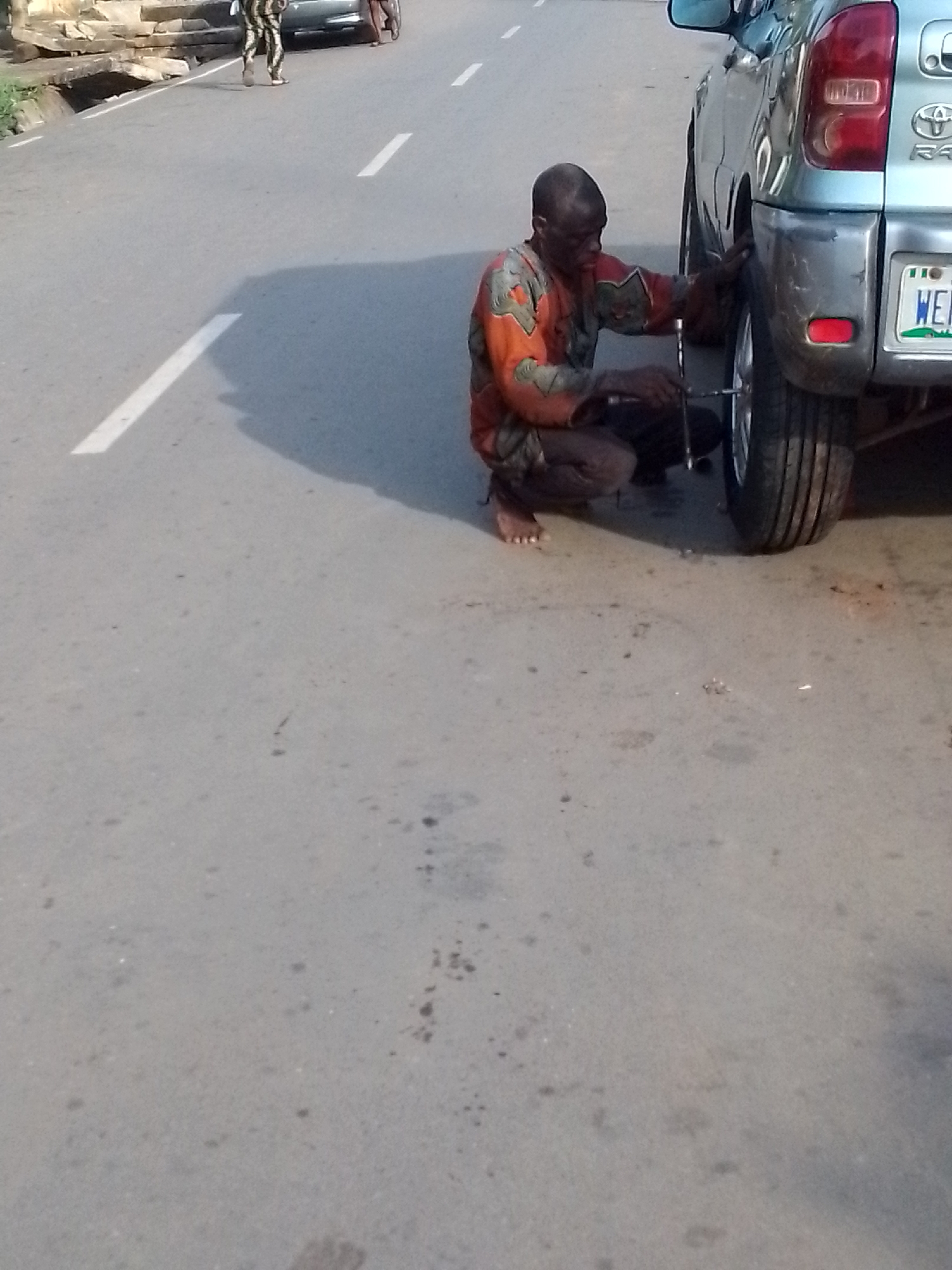 The image shows a man kneeling next to a silver Toyota RAV4 on a paved road in Orlu, Nigeria. The man is wearing an orange and brown patterned shirt and dark pants. He is using a lug wrench to work on the front passenger side tire of the vehicle. The car is parked on the right side of the road, with a white line marking the lane. There is another person walking in the background, on the opposite side of the road. The road appears to be in a residential or commercial area. The car has a license plate, which reads "WE". The time of day appears to be daylight. There are scattered small dark stains on the road surface.