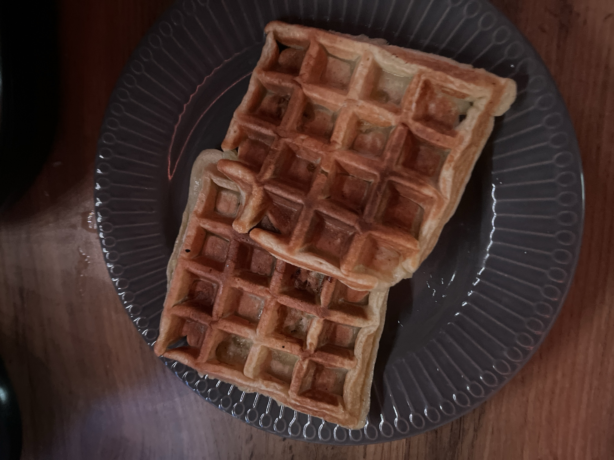Two golden-brown square waffles sit on a grey plate with a decorative edge. The plate rests on a wooden surface. The location is not identifiable from the image.