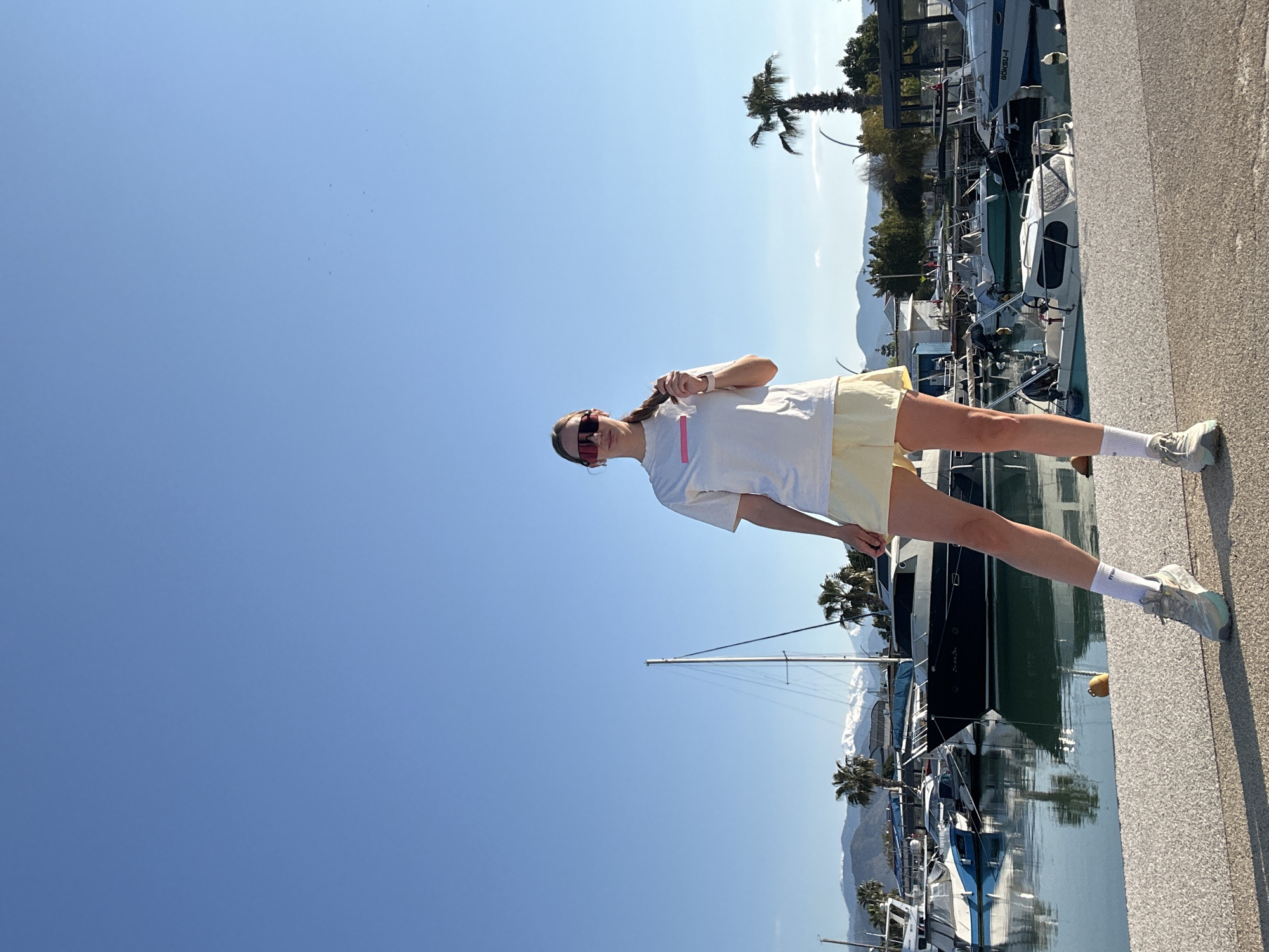 A medium, outdoor shot shows a woman standing on a concrete pier in Fethiye, Turkey. She faces forward and slightly left, with a braid in her hair, sunglasses, a white t-shirt with a pink stripe, and yellow shorts. She is holding her braid. The pier is to the right of the frame. Behind her, there is a harbor with many boats docked. In the background, there are palm trees and mountains. The sky is clear and blue.