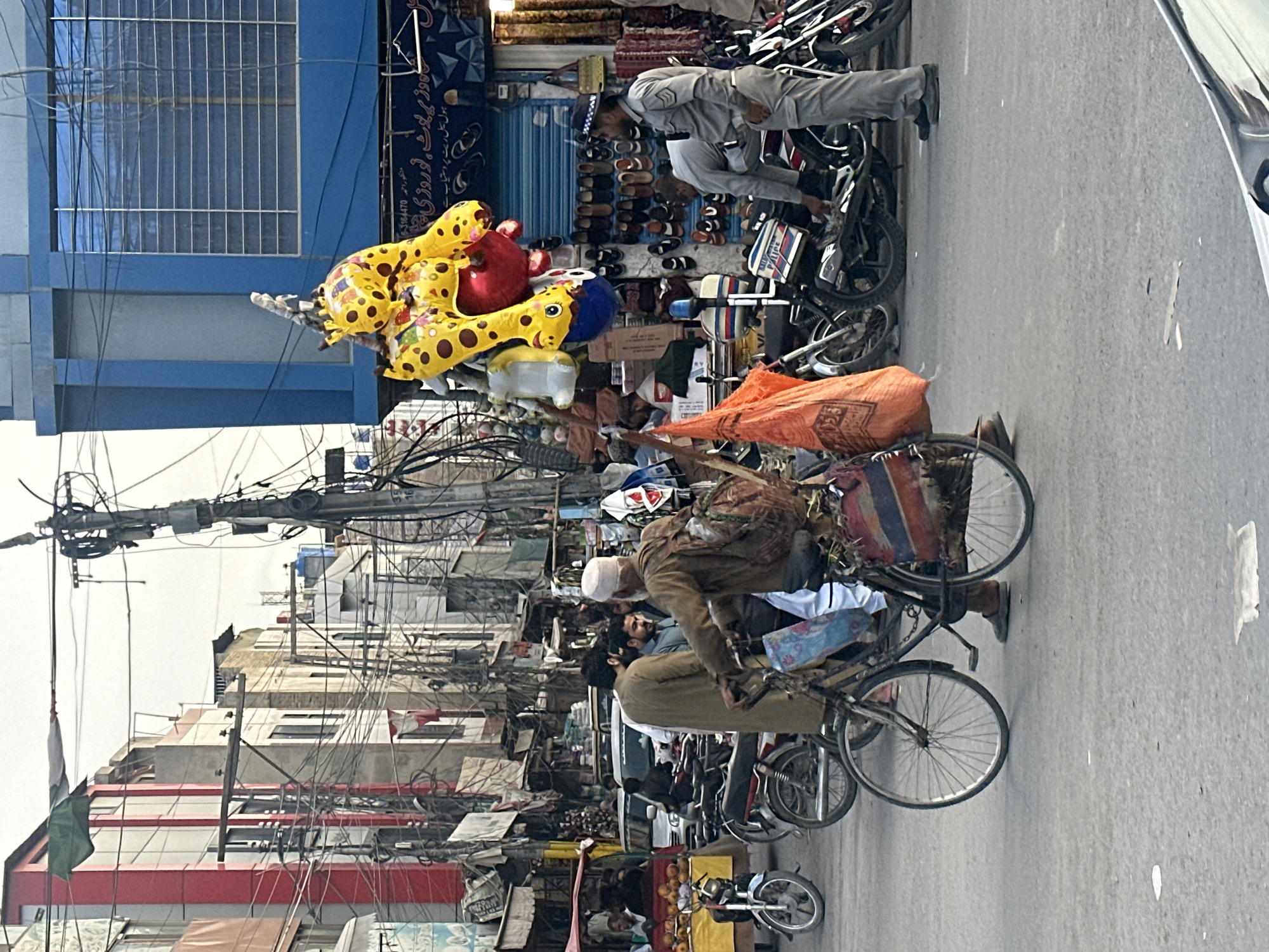 The image depicts a busy street in Quetta City Tehsil, Pakistan. Several people are seen riding bicycles laden with goods. One vendor carries a large bunch of balloons, while others have bags and various items strapped to their bikes. A police officer is inspecting a motorcycle parked along the side of the street. Shops line the street, displaying footwear and other merchandise. Overhead, a chaotic network of electrical wires is visible against the backdrop of buildings.