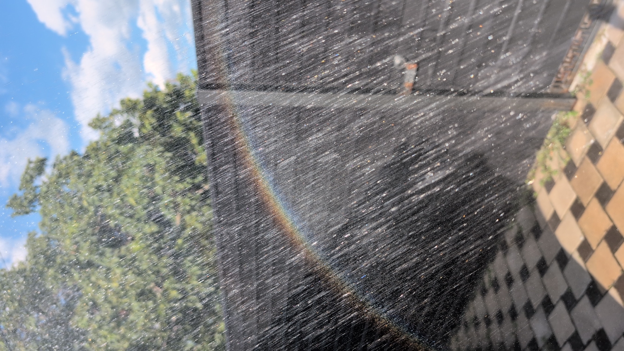 A faint rainbow arcs across a dark, slatted fence as water streams down from above, creating a shimmering effect against the fence's surface. The fence is partially obscured by the falling water, with a metal latch visible on the right side. Below the fence, a patterned patio of interlocking square tiles, alternating in dark and light shades, is visible. Lush green foliage can be seen in the background, with the sky above featuring patches of blue and white clouds. The image appears to capture a moment during a light rainfall or sprinkler activity on a sunny day, suggesting an outdoor residential setting in Izmail, Ukraine. There are no people present in the image.