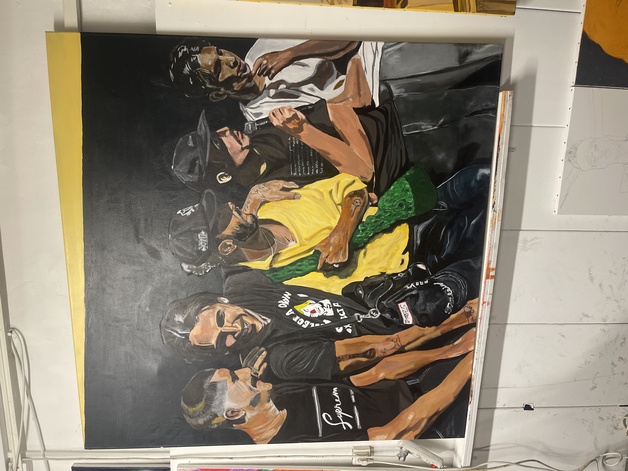 In a studio or workshop located in Marseille, France, a large, rectangular painting of five young adult males is displayed, leaning against a white, vertically paneled wall. The painting itself is approximately 1.8 meters tall and 1.2 meters wide, featuring the figures from the mid-torso up, set against a dark, brushed background with a horizontal yellow stripe spanning the top edge. From left to right within the painting: 1. **First Figure:** A light-skinned male with short, faded hair and a crew cut, wearing a black t-shirt with a white "Supreme" logo. He is depicted in profile, laughing heartily, looking towards the second figure. His right arm is bent at the elbow. 2. **Second Figure:** A light-skinned male with dark, wavy hair, also laughing and looking towards the first figure. He wears a black t-shirt with a graphic design of a clown's head and the text "COLLECT A OWN". A black messenger bag, detailed with a white "Titleist" tag and "PROVOK" lettering, is slung across his chest. His left arm is draped over the shoulder of the first figure. A black tattoo is visible on his right forearm. 3. **Third Figure:** A male with medium-dark skin tone, a dark beard, and a mustache, wearing a black baseball cap. He wears a bright yellow long-sleeved shirt. A textured, green, possibly knitted or woven item is draped over his left arm, which rests on the shoulder of the fourth figure. His right hand holds a small, dark, indeterminate object. He looks downwards and to his right. 4. **Fourth Figure:** A light-skinned male wearing a dark baseball cap and a dark t-shirt with faint, light-colored script. He holds a black microphone in his right hand, positioned near his mouth, indicating speech or singing. His expression is focused or serious, and he looks towards the right. 5. **Fifth Figure:** A male with medium-dark skin tone and dark, swept-back hair, wearing a white t-shirt and grey trousers. His left hand is raised to his chin, suggesting a contemplative or pensive demeanor. He looks downwards and to his right. The painting rests on a white wooden ledge or shelf, which is visibly splattered with various colors of paint. The white paneled wall behind the painting exhibits minor scuffs and a small, dark, rectangular indentation on its left side. Above the painting, white electrical wires are secured along the wall with clips. To the left, a white power