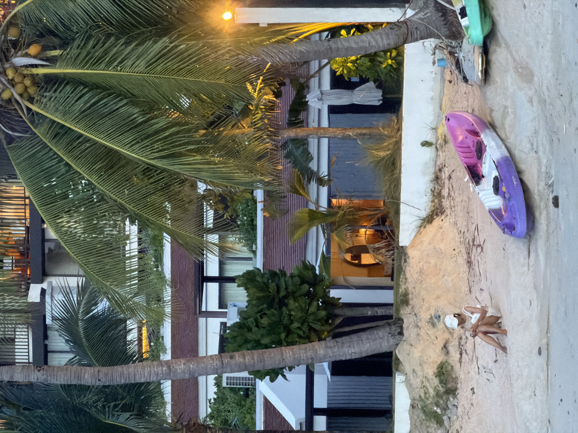 The image depicts a tropical beach scene at dusk near Baan Bang Rak, Thailand. A woman wearing a white hat and white attire is seated on the sand, looking down at her phone. In the foreground, a purple and white kayak rests on the beach, with another kayak partially visible to its right. The background features a multi-story building with balconies and palm trees. A bright light fixture illuminates the scene from the right side of the image. The sand is light brown and textured, with scattered debris.