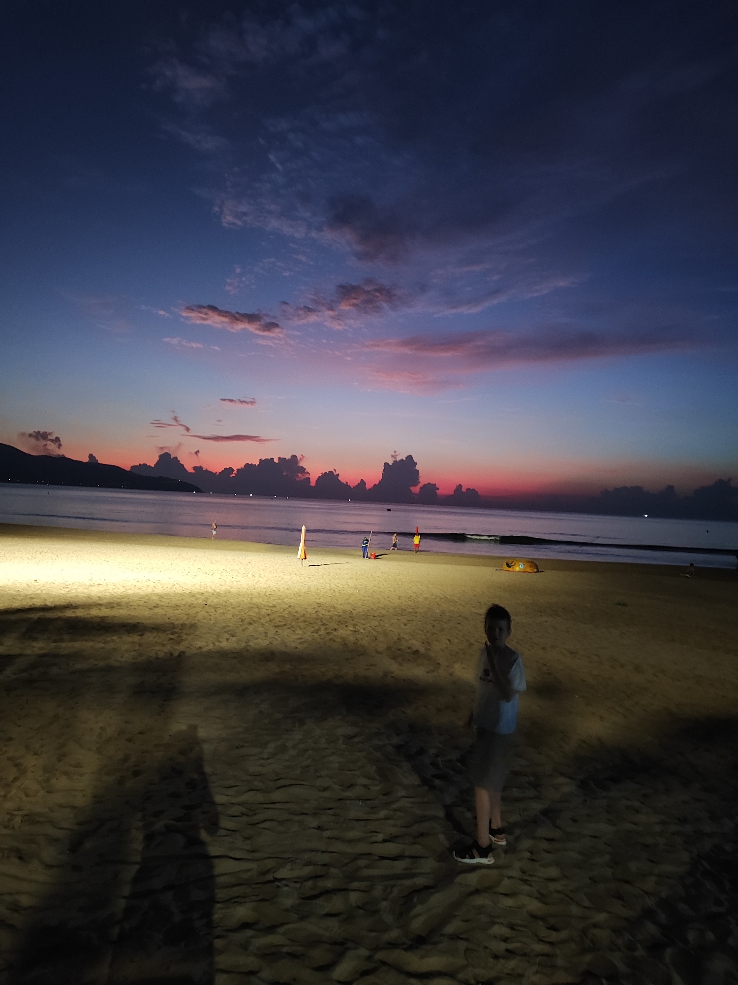 A wide-angle shot of a beach at dusk in Đà Nẵng, Vietnam. A boy stands in the foreground on a sandy beach, casting a long shadow. In the middle ground, the beach is illuminated, revealing beach umbrellas and figures of people further out. The sea is calm, with a line of dark mountains silhouetted against the horizon. Above the mountains, the sky transitions from a dark blue at the top to pink and red near the horizon. Clouds are scattered across the sky.