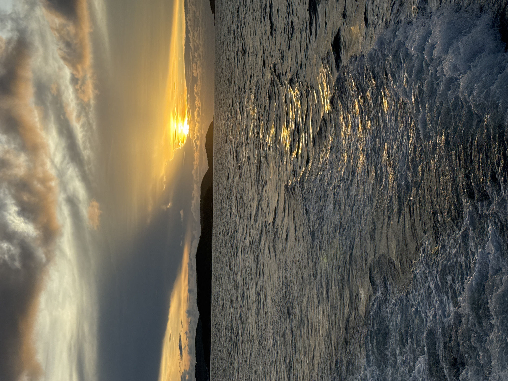 The image depicts a scene at sunset in Pattaya City, Thailand. The left side showcases the sky with a blend of dark and light clouds tinged with gold, indicating the setting sun which is partially obscured by the clouds and a dark landmass. The sun's rays peek through, creating a bright spot. The right side displays a body of water reflecting the golden light, with visible wave patterns and varying shades of gray and white foam. The image has been rotated.