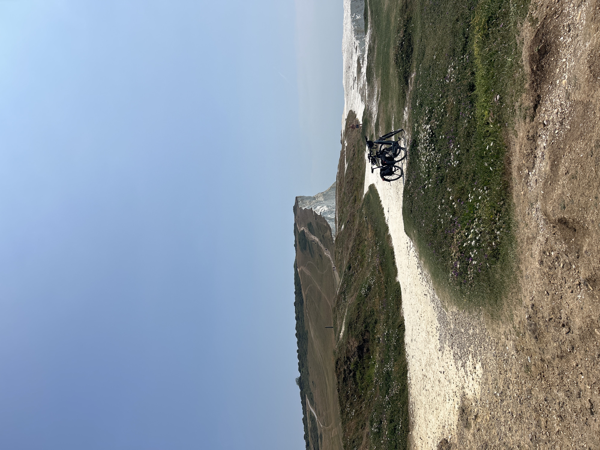 The image depicts a scenic view of the Seven Sisters cliffs in Seaford, United Kingdom, on a clear day. In the foreground, a dirt path leads to a white gravel path that winds through lush green grass dotted with wildflowers. Two bicycles are parked on the gravel path, facing towards the sea. In the middle ground, a lone figure stands near the edge of the cliff, looking out at the ocean. The iconic white chalk cliffs of the Seven Sisters stretch across the horizon, with walking paths visible on their slopes. The vast expanse of the blue sky dominates the upper portion of the image.