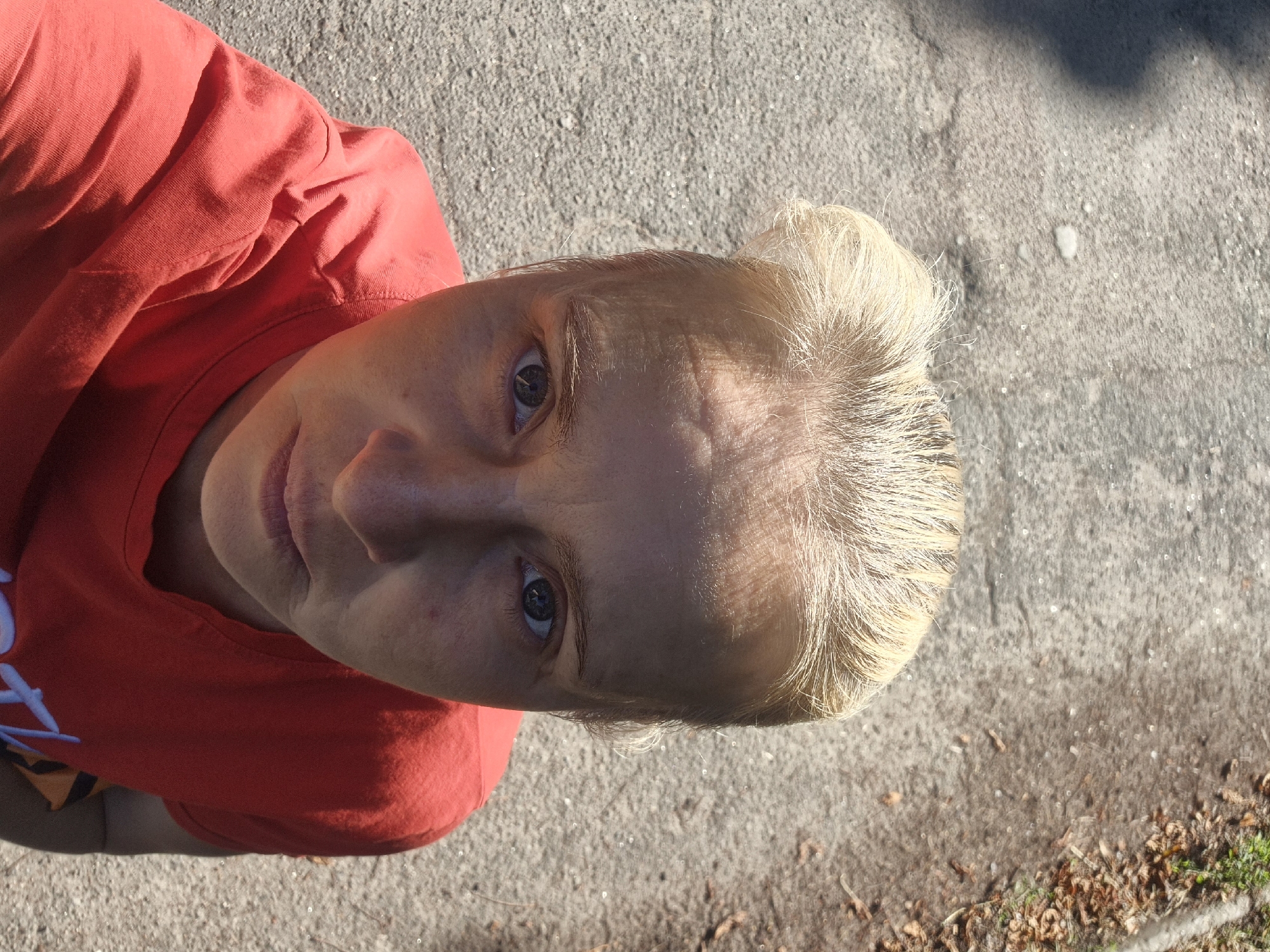 The image is a high-angle, close-up, selfie-style shot of a fair-skinned woman with blonde hair pulled back. She is wearing a red t-shirt. Her face is partially illuminated by sunlight, casting shadows. The background is an asphalt surface, appearing to be a road or path, with scattered leaves and some green foliage visible on the right side. The setting is consistent with an outdoor location in or around Sozonivka, Ukraine, likely during the daytime. The focus is on the woman's upper body and face.