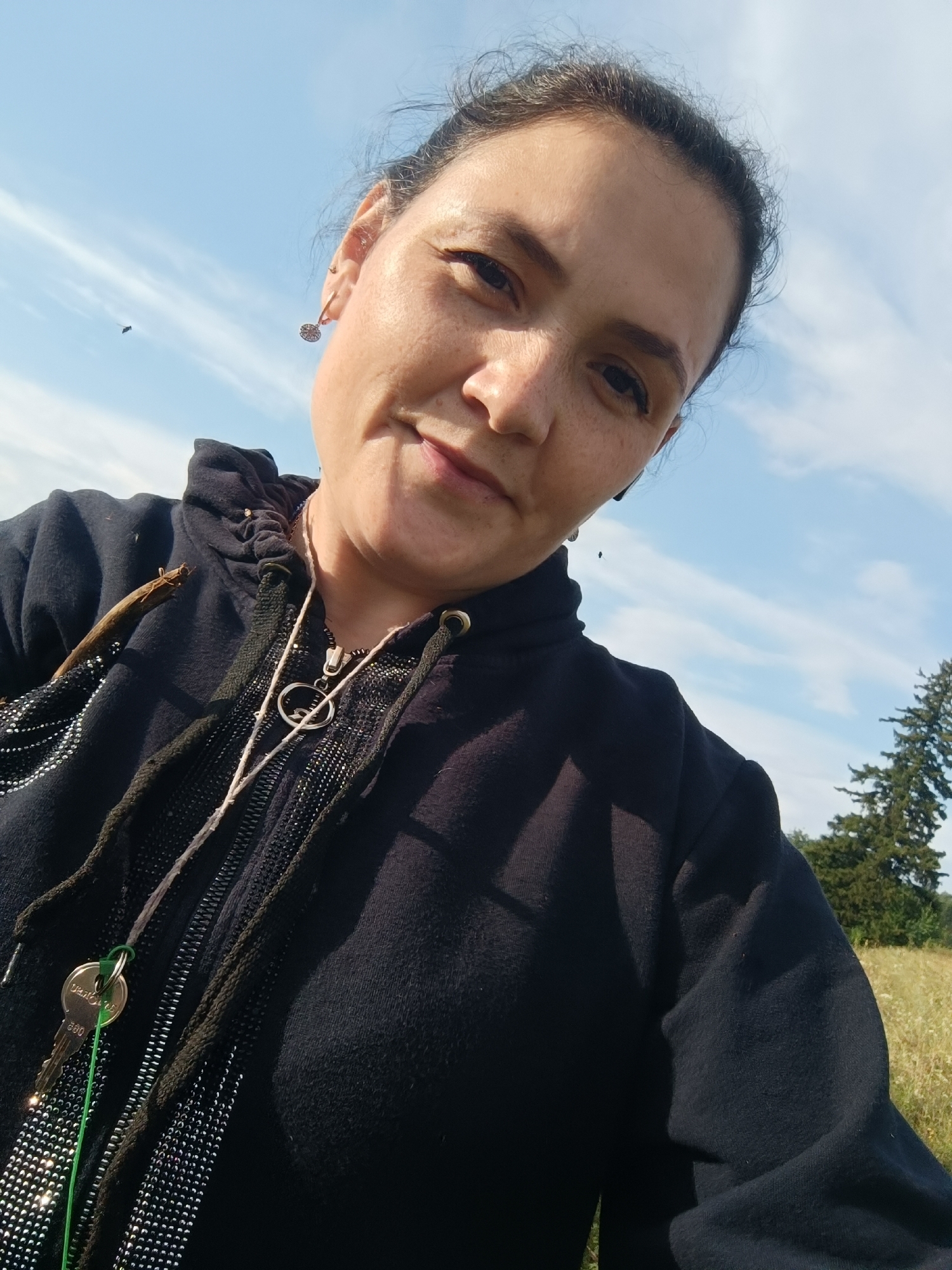A woman is posing for a selfie outdoors under a blue sky with some clouds. She has dark hair pulled back and is wearing a black hooded sweatshirt. She is wearing earrings and a necklace with a circular pendant and a key attached. The background consists of a grassy field and some trees. The scene appears to be a rural or park area. There is no indication of the location being Karsha, Russia.