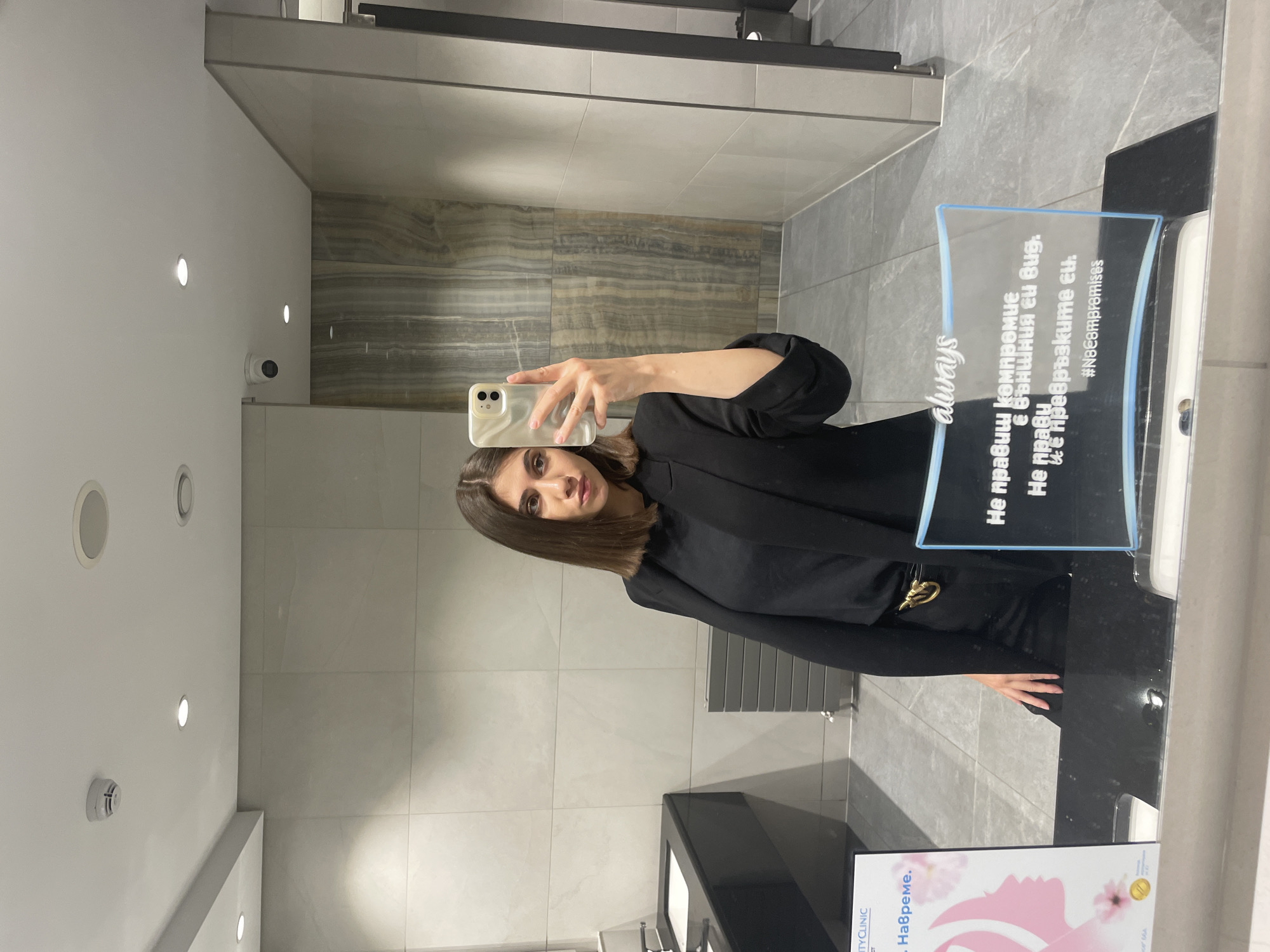 Here is a description of the image.
The image is a selfie taken in a bathroom, likely in a commercial or public setting. The subject, a woman with short, dark hair, is holding her phone to capture the photo in the mirror. She is wearing a black outfit and has a gold-toned accessory at her waist.
The bathroom has a modern design with grey tiling on the walls. There are recessed lighting fixtures in the ceiling. In the mirror's reflection, there is a marble-like wall behind the woman, as well as a radiator and a sink area with a black countertop.
A sign is placed near the mirror with text written in Bulgarian, promoting something along the lines of "beauty clinic." The sign also has the word "always" written in English and a hashtag: #NoCompromises. The text includes phrases such as "Не правиш компромис е външния си вид."