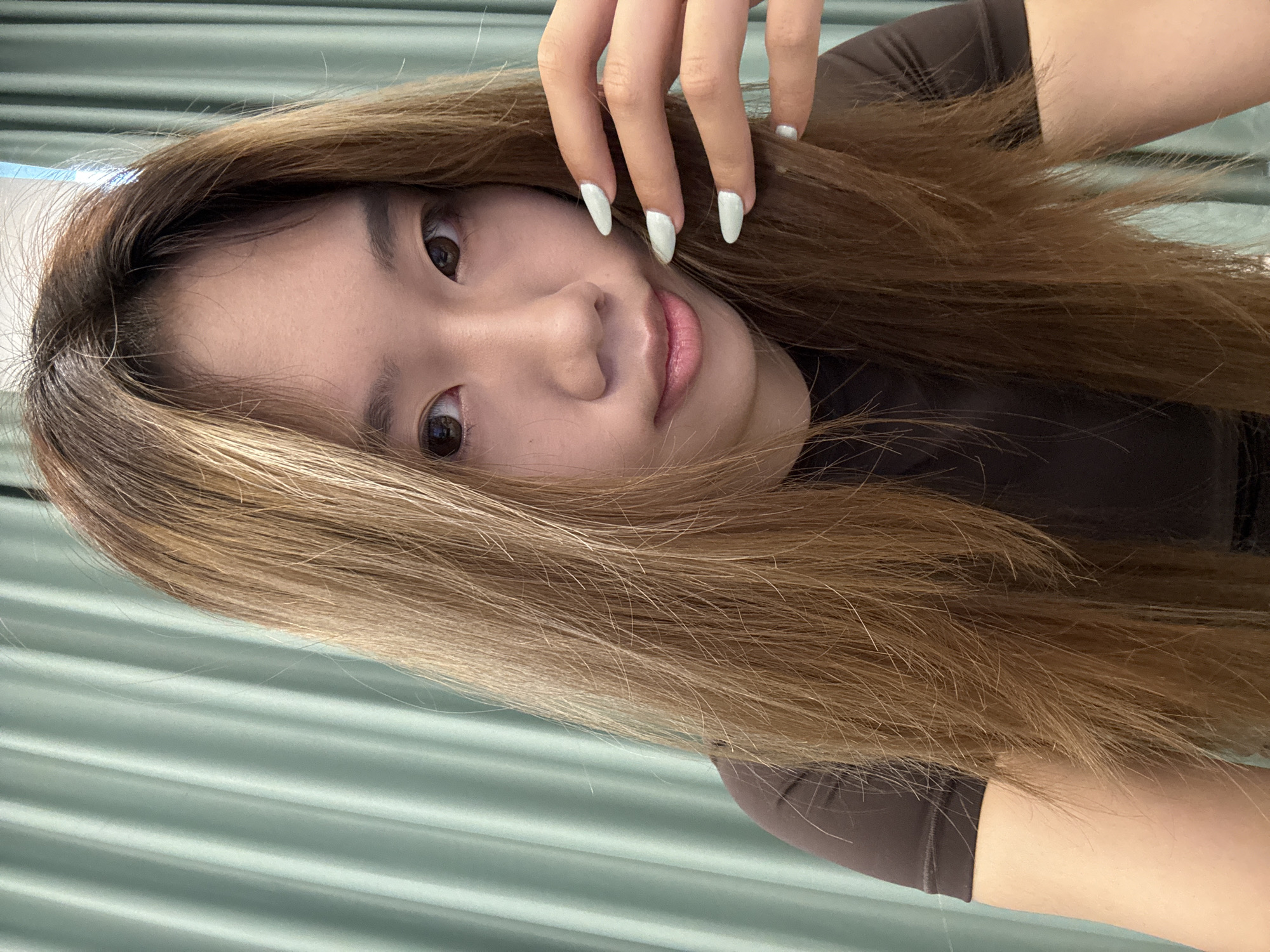 A young woman is pictured indoors, likely in Nan'an District, China, although specific location details are not evident from the image. She has brown hair with lighter highlights and is wearing a dark brown top. Her left hand is raised near her face, displaying long, white fingernails. The background appears to be a light green, ridged surface, possibly a wall or blinds.