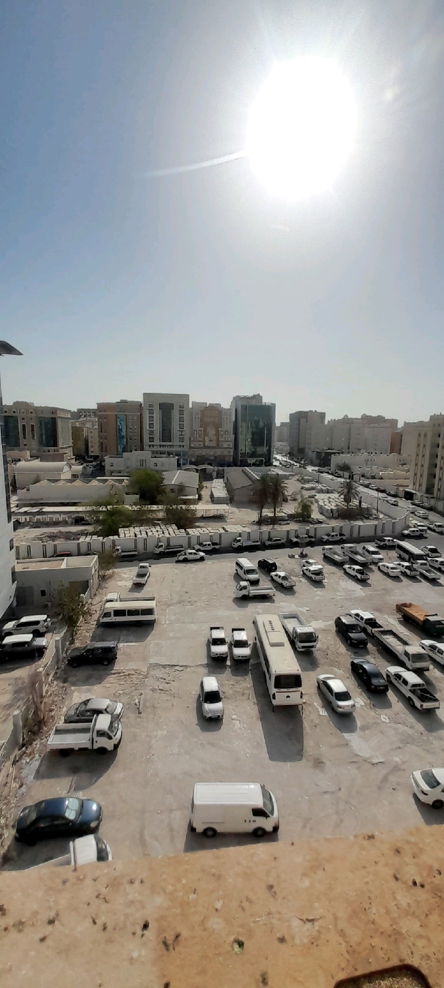 A high-angle, full-shot, vertical outdoor photograph captures a sun-drenched cityscape in Doha, Qatar. The bright, diffused sun dominates the upper portion of the frame, casting a lens flare and a faint contrail across the clear, pale blue sky. Below, a dense urban landscape unfolds, characterized by a collection of modern, multi-story buildings of varying architectural styles, predominantly featuring light-colored facades and reflective glass windows.
The lower half of the image reveals a vast, paved outdoor parking lot filled with numerous vehicles. These include white and dark-colored cars, several white vans, and at least one articulated bus. A few pickup trucks, some with open beds, are also visible. The parking lot appears to be adjacent to a series of low-rise structures, possibly commercial or industrial buildings, some of which have corrugated metal roofs. Evidence of ongoing construction or storage is present with stacks of materials or modular units visible in some areas. Sparse vegetation, including a few palm trees, dots the scene, hinting at a warm climate. The ground surface of the parking lot is a mix of asphalt and exposed, rough soil or gravel, with patches of lighter-colored concrete. Shadows are cast by the buildings and vehicles, indicating the time of day is likely late morning or early afternoon. There are no visible people in the frame.