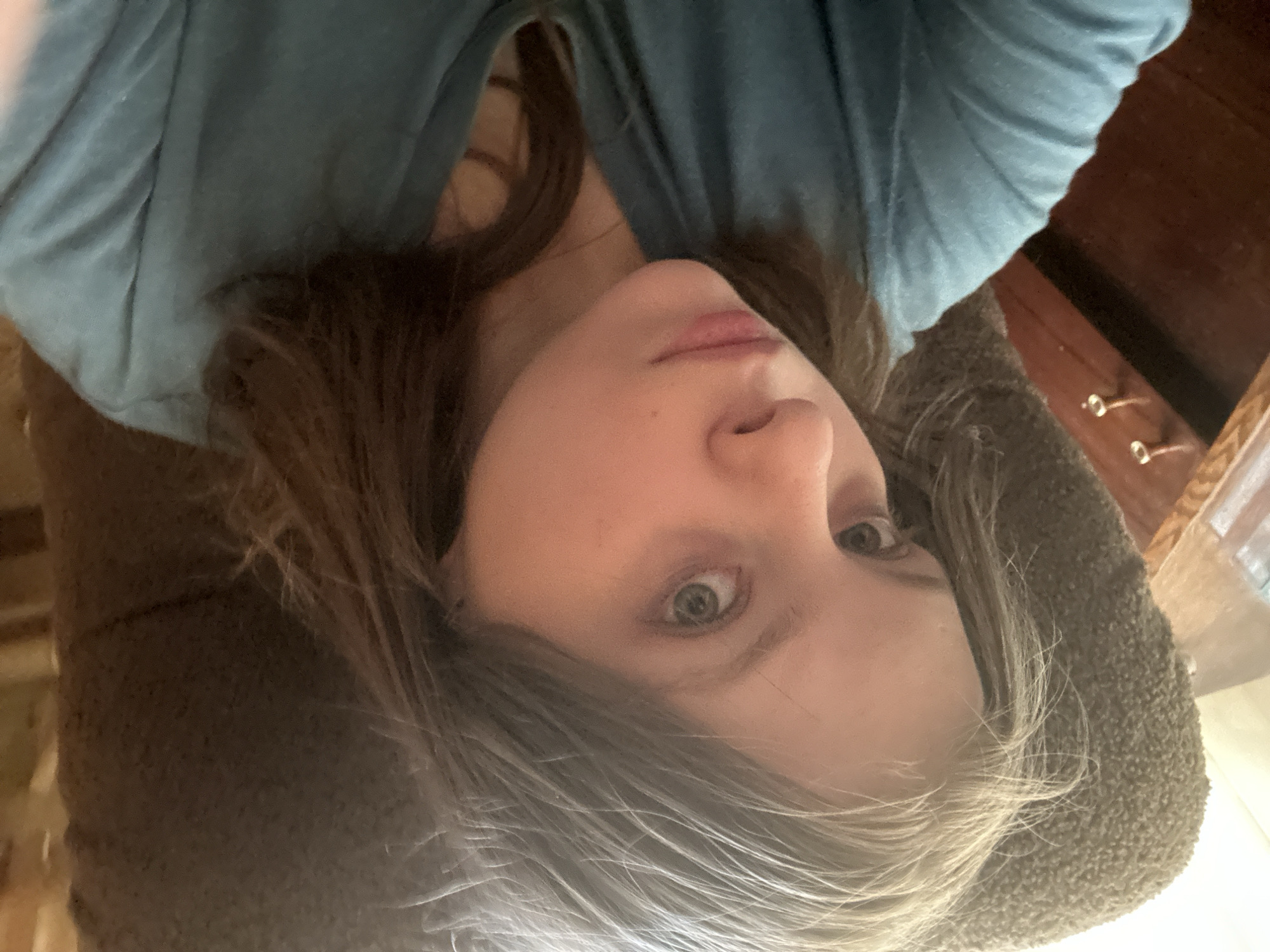 A close-up selfie of a person with fair skin, grey eyes, and brown and grey hair. The person is wearing a blue shirt. The background includes a brown upholstered object, possibly a chair or couch, and a dark brown wooden surface with drawer pulls, possibly a table or dresser. There is no indication of the location being Moscow, Russia.