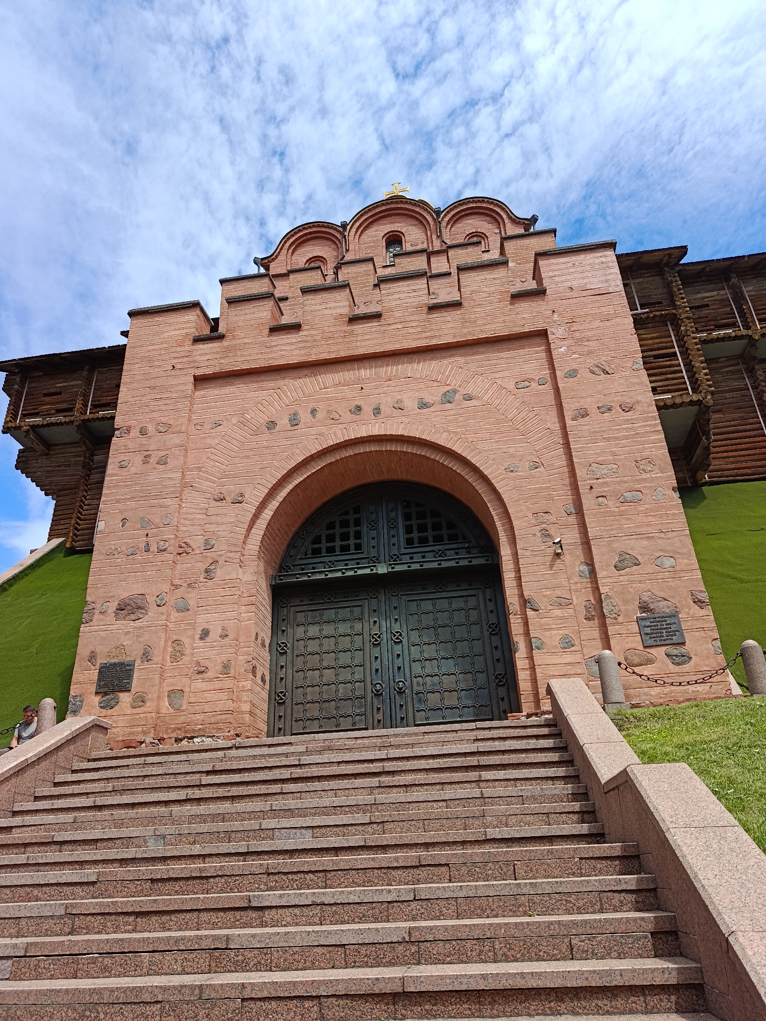 The image shows the Golden Gate of Kyiv, Ukraine, a historical monument. A wide, grand staircase made of pink granite leads up to the main entrance of the gate. The gate itself is constructed of red brick with stone accents and features a large, ornate metal door. Above the entrance, the gate has a crenellated design and decorative arched elements, topped with a golden cross. To the right and left of the gate, wooden structures are visible, likely reconstructions of historical fortifications. The area around the gate is covered in vibrant green grass. The sky above is a bright blue with scattered white clouds. A single person is visible sitting on the left side of the staircase. The overall scene is captured from a low-angle perspective, emphasizing the grandeur of the monument.
