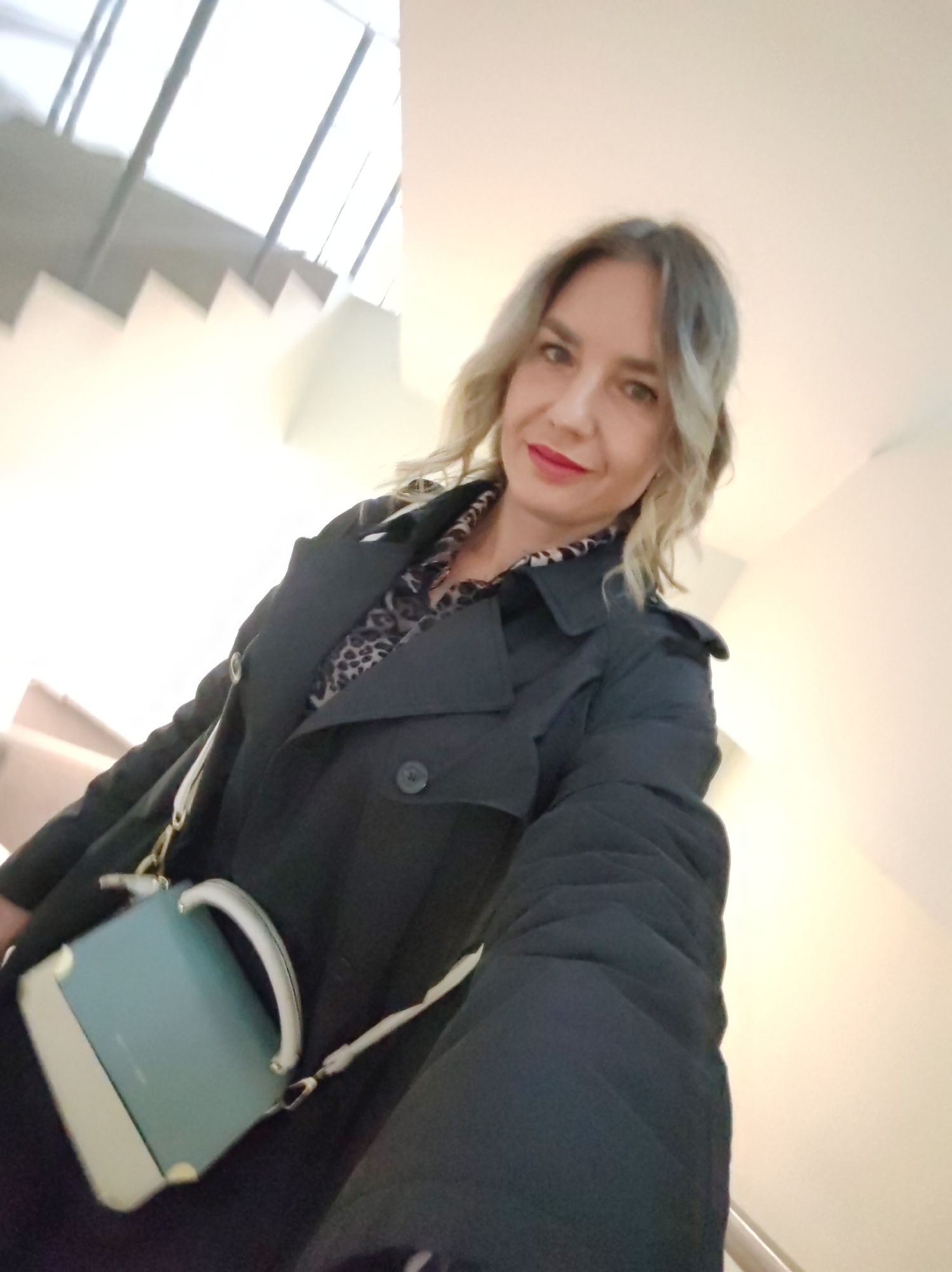 A woman poses for a selfie indoors in Petropavlovsk-Kamchatsky, Russia. She has fair skin, wavy light brown hair with blonde highlights, and wears red lipstick, looking directly at the camera with a subtle smile. She is dressed in a dark, possibly black, trench coat or long jacket, with a leopard-print shirt visible underneath. A small, structured handbag, light blue-green with cream accents and gold-toned hardware, hangs across her body. The background features a brightly lit, modern interior with white walls and a white staircase ascending upwards. Reflections of windows or skylights are visible in the upper part of the image, indicating a well-lit space. The photo is taken from a slightly low angle, suggesting the woman is standing on a lower step or at the base of the stairs. There is no visible text.