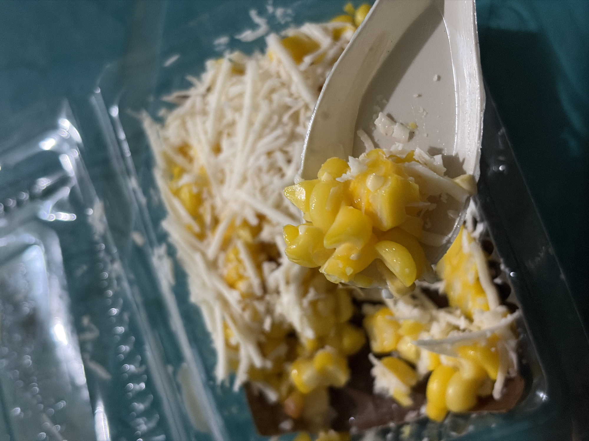An overhead close-up shot shows a plastic container of corn kernels mixed with shredded cheese. A white plastic spoon is lifted, carrying a scoop of the corn and cheese mixture. The corn is bright yellow, and the cheese is finely shredded and white. The container is clear plastic, and the background is a blurred teal color, possibly a table. There is no indication of the city of Tupah, Indonesia, or any other specific location.