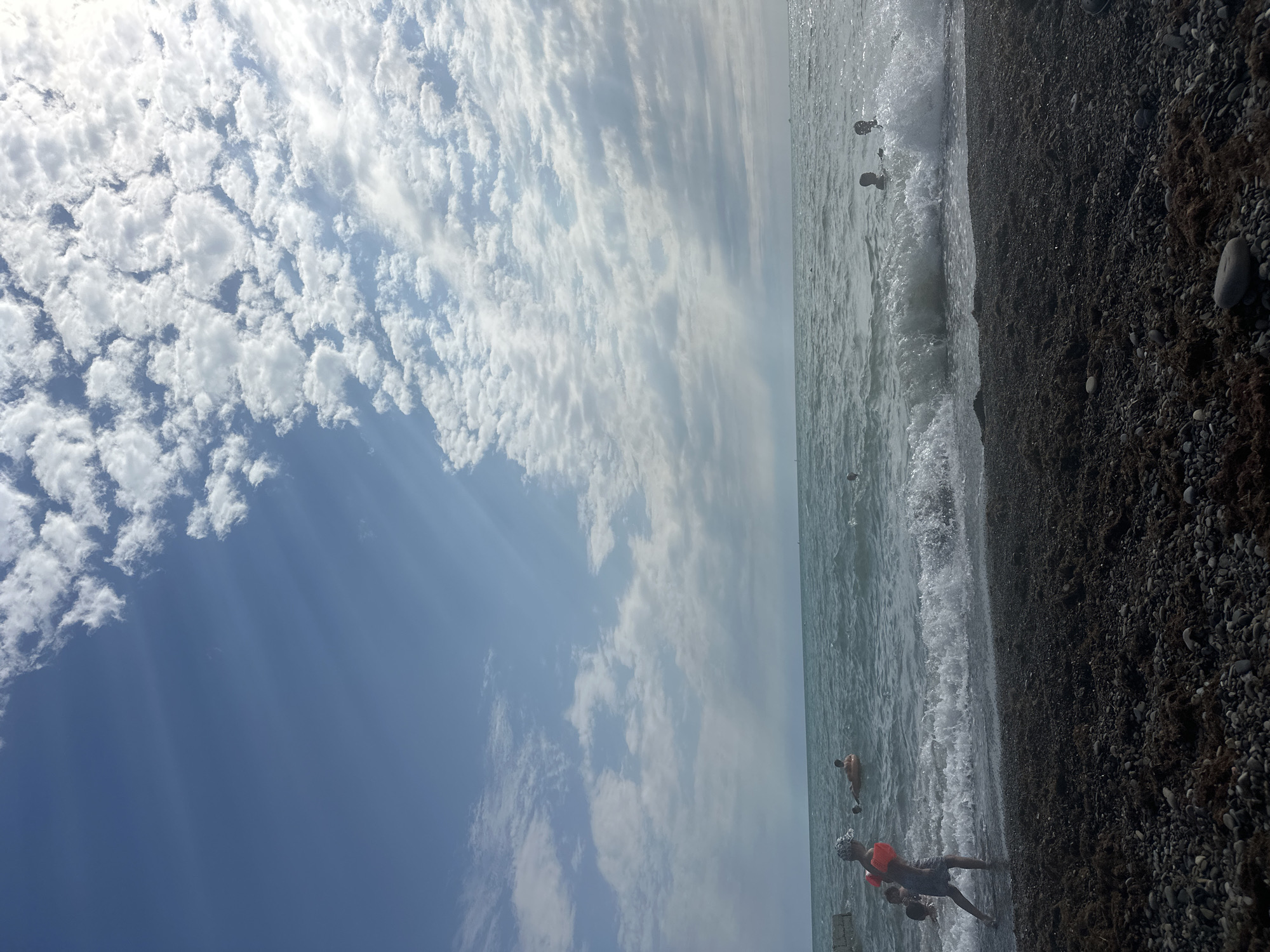 A beach in Khostinskiy, Russia, features a mix of dark pebbles and organic matter on the shoreline. The sea is a choppy, teal color with white-capped waves breaking near the shore. In the water, several people are visible. One individual, wearing an orange floatation device, walks towards the water from the left side of the frame. Another person floats on an orange inflatable ring further out in the sea. Other figures can be seen swimming or playing in the water. The sky above is a bright blue, with scattered, fluffy white clouds that are illuminated by sunlight, creating streaks of light that descend towards the sea. The overall atmosphere is one of a warm, sunny day at the beach.