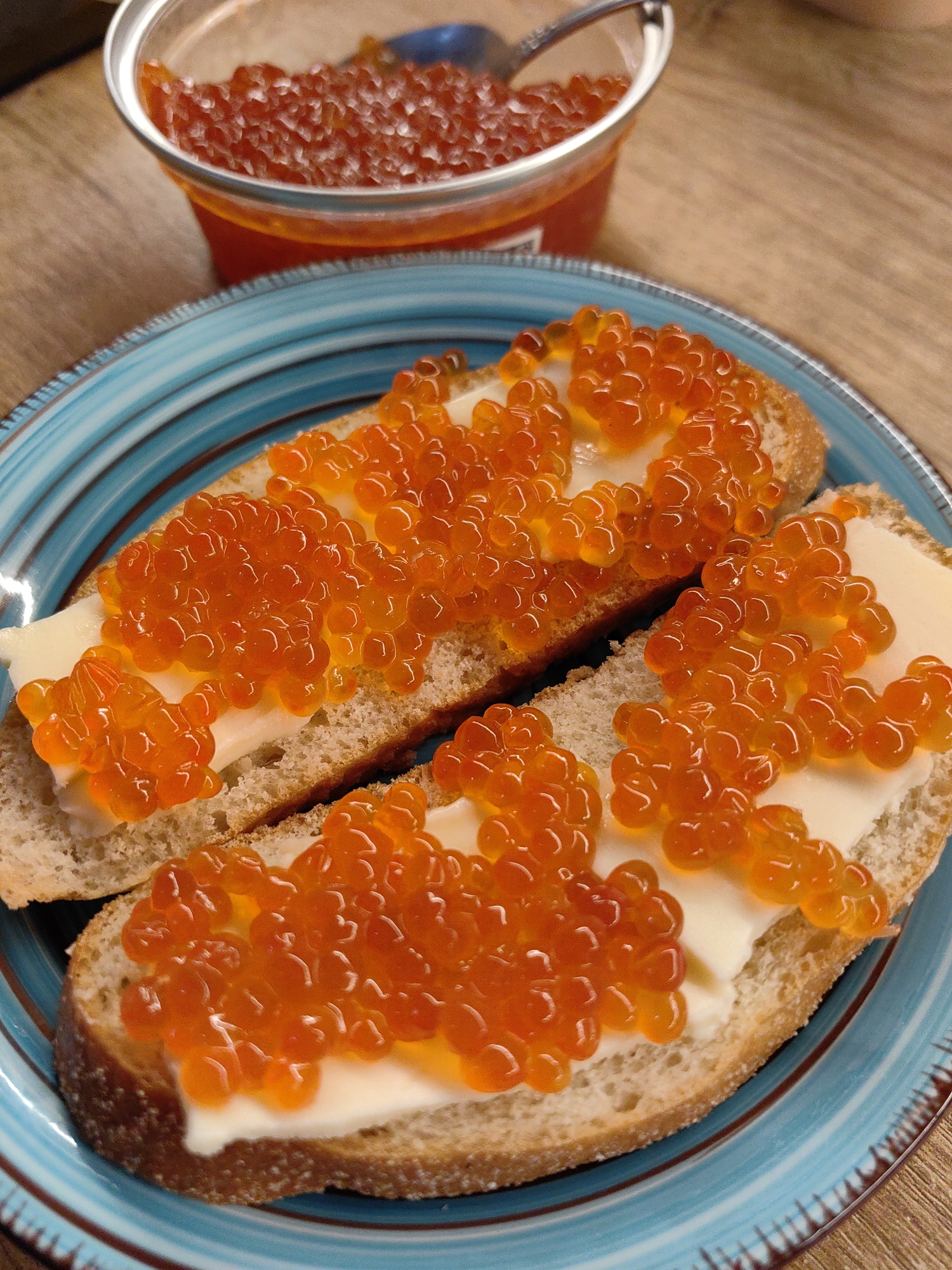 Two slices of light brown bread, spread with a thin layer of off-white butter and generously topped with glistening red caviar, are positioned on a round, textured blue ceramic plate with a brown rim. The plate occupies the lower two-thirds of the vertical frame. In the upper-left background, a clear plastic container filled with additional red caviar is visible, with a small silver spoon resting within it. The items are placed on a light-brown wooden surface, possibly a table, exhibiting a visible grain pattern. No people are visible. The location, including a specific city like Sochi, Russia, cannot be determined from the visual information provided in the image.