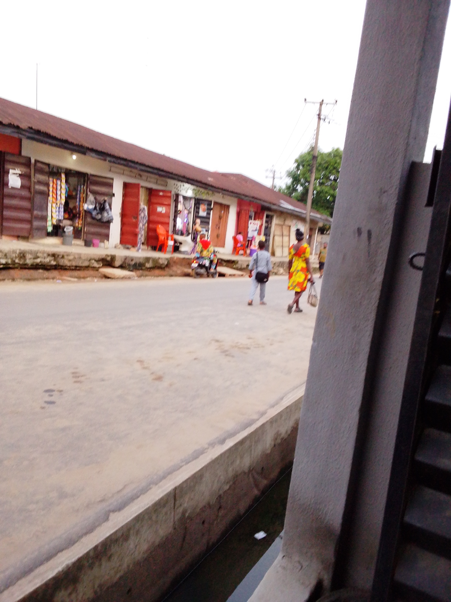 A street scene in Aba, Nigeria, is presented from a high-angle vantage point, partially obscured by a concrete structure. The street has a low-lying concrete barrier along its edge, concealing a drainage channel. A row of single-story buildings lines the street, their facades painted in alternating colors of red, brown, and white, with corrugated metal roofs. Shops along the street are visible, with goods displayed in front.
Several individuals are walking along the street, including a woman in a yellow and orange dress, a man in a grey shirt and pants, and a motorcyclist wearing a red helmet and vest. A utility pole is visible in the background, and trees line the street farther down.
