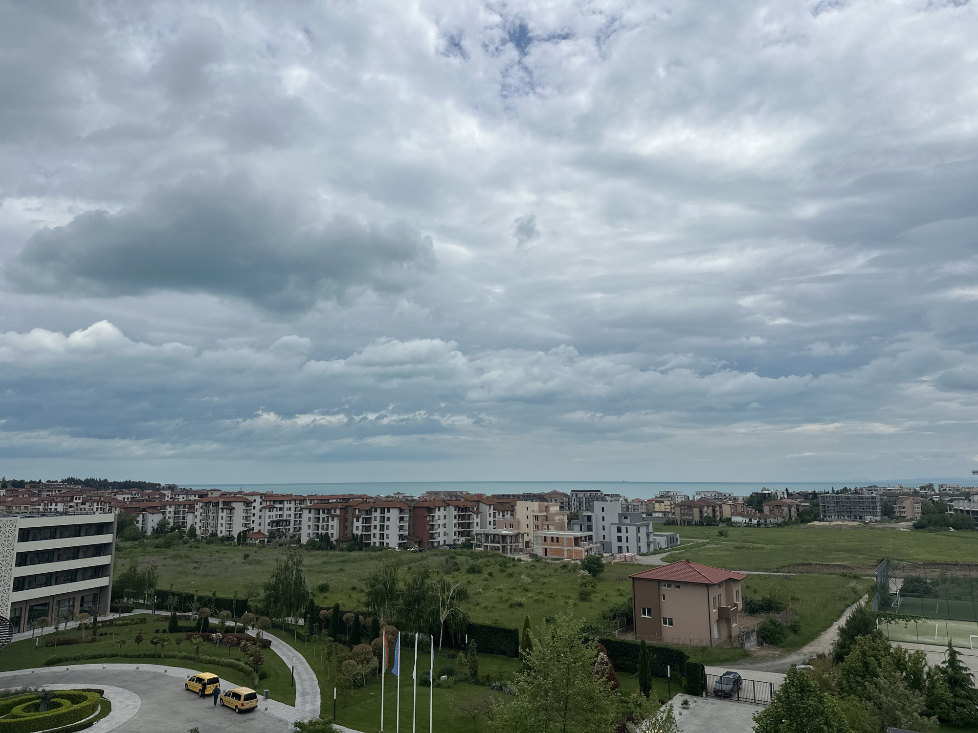 The image shows a landscape in Ravda, Bulgaria under a cloudy sky. The sea is visible in the distance, behind a line of buildings with red tile roofs. Closer to the viewer, there's a green space with scattered trees, small bushes, and landscaped lawns. There are a few yellow vehicles parked in a circular driveway and a tennis court. Flags are flying. A single-story house with a red tile roof is also visible. The scene appears still and unoccupied.