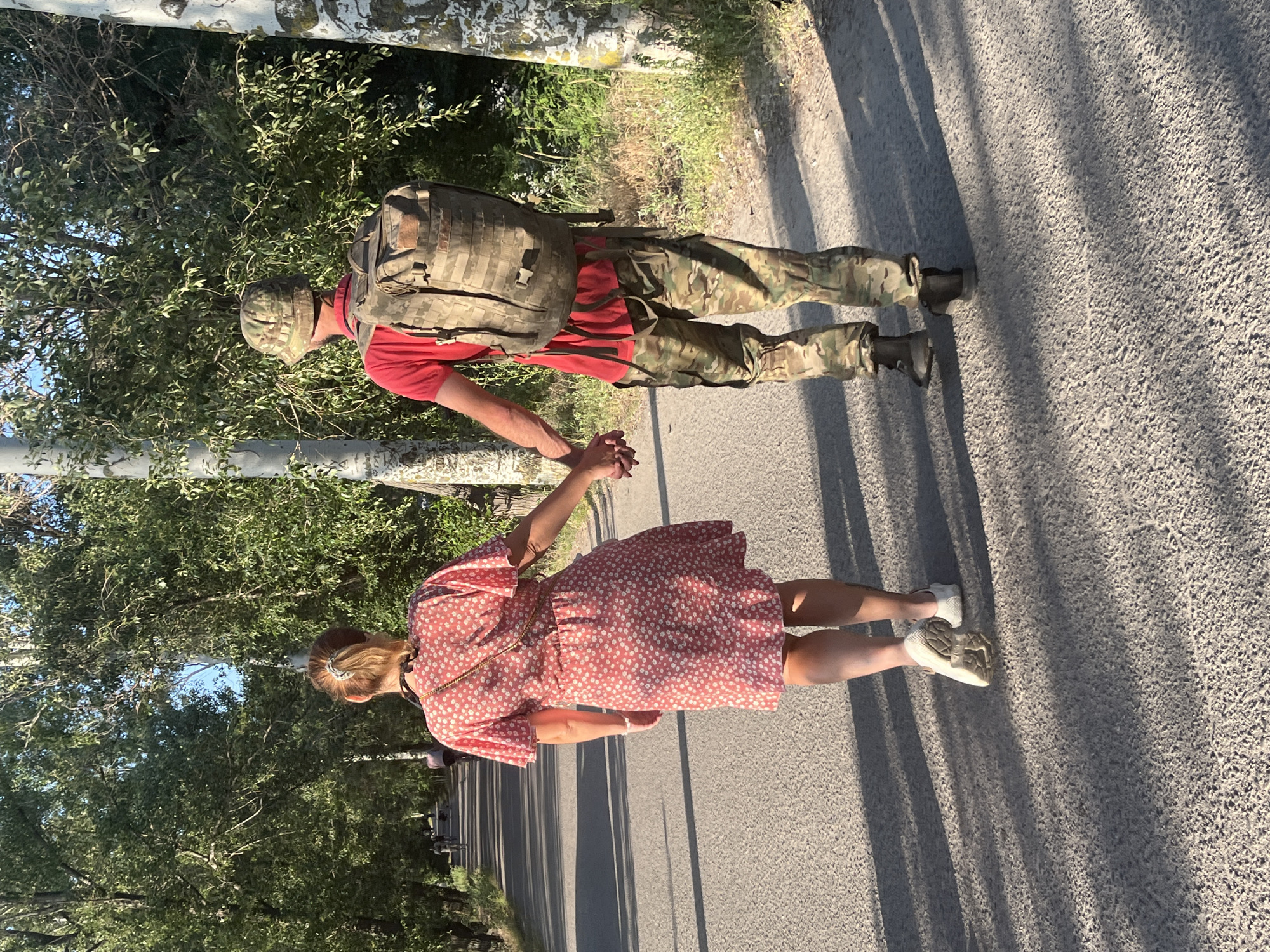 Two people, a woman in a pink floral dress and a man in camouflage pants, a red t-shirt, and a camouflage hat, are walking hand-in-hand on an asphalt path lined with trees. The man is wearing a large backpack. The path appears to be in a park or a tree-lined avenue. The scene is bathed in sunlight, casting long shadows on the path. In the background, other people are visible in the distance, some appearing to be on bicycles. The environment suggests an outdoor recreational area within a city, possibly Dnipro, Ukraine, given the context provided.
