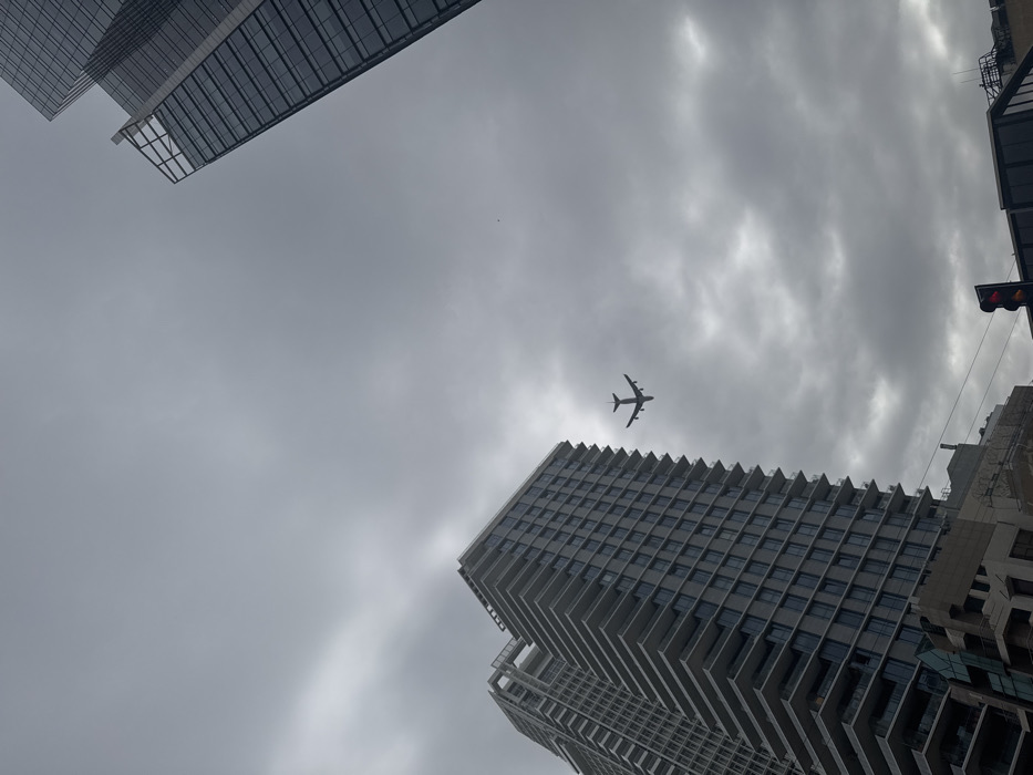 The image displays an urban scene in Tel-Aviv, Israel, under a predominantly overcast, grey sky. A large, dark-silhouetted passenger aircraft, consistent with a wide-body, four-engine jet, is prominently featured in the upper-middle of the frame, flying horizontally with its underside visible against the clouds.
On the left side of the frame, extending from the bottom-left, stands a tall, modern residential or office building. It features a grey, stepped facade with numerous balconies and dark-tinted windows, characteristic of luxury high-rise architecture found in areas such as Park Tzameret in northern Tel-Aviv. Another similar, but less detailed, building is partially visible behind it. On the far right, a sleek, dark-glass skyscraper is partially visible, reflecting the cloudy sky.
At the bottom-right, a traffic light with a red illuminated signal is visible, indicating a street intersection below the vantage point of the photograph. Utility lines or cables are faintly discernible crossing the lower portion of the image. No individuals are visible in the scene. The overall lighting is diffused, consistent with an overcast day, highlighting the dark silhouettes of the aircraft and elements of the cityscape.