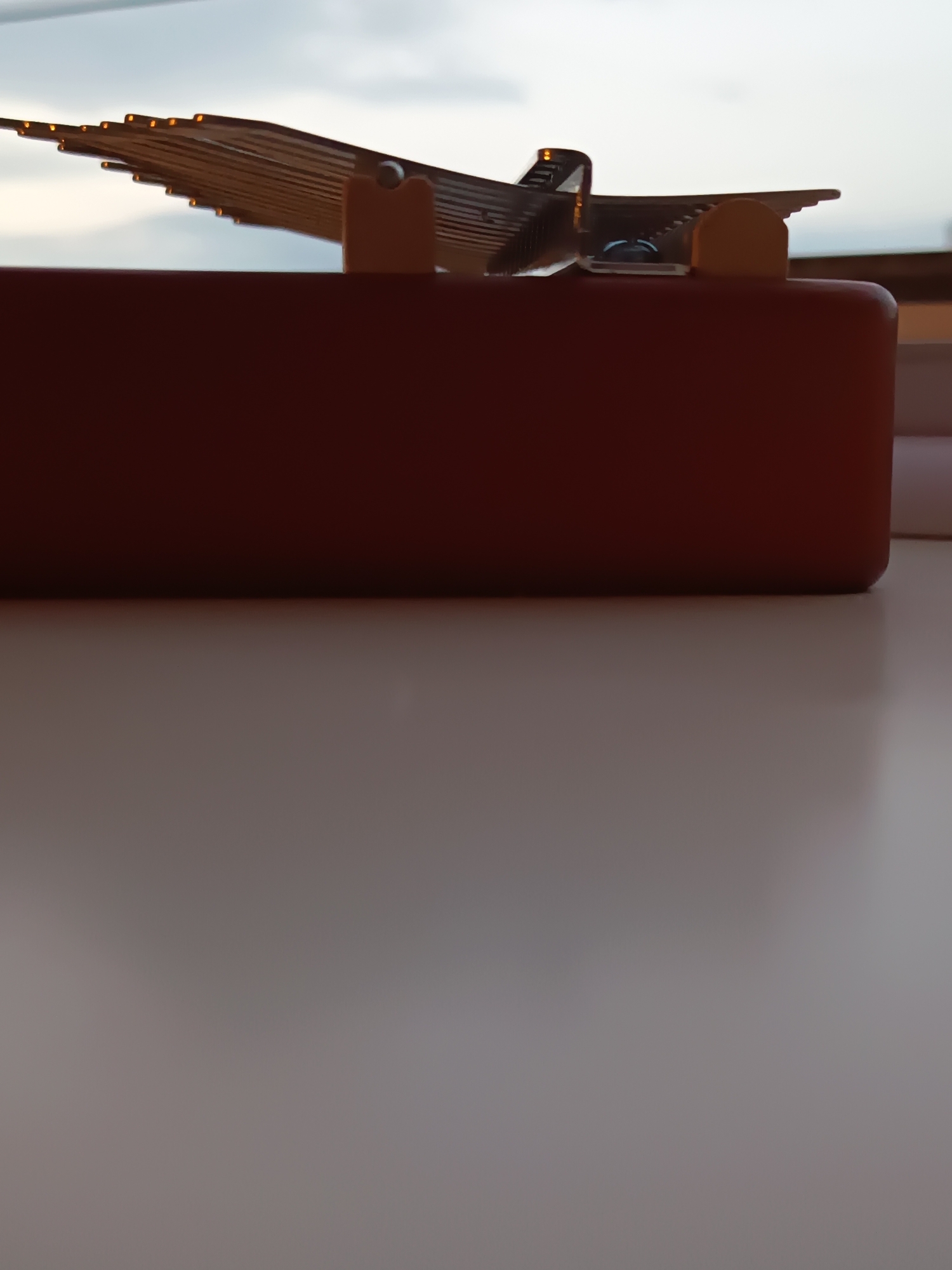 The image displays a close-up, low-angle shot of a brown rectangular object, possibly a box or a lid, positioned horizontally. On top of this object, a metallic structure with multiple thin, angled slats resembling a wing or a fan is visible, with small illuminated elements at the edge of some slats. The metallic structure is supported by a small, curved, orange-colored component. The background consists of a blurred sky with clouds. The scene is indoors, likely on a surface like a table, with a reflective quality, casting a faint shadow of the brown object. There are no people or discernible interactions depicted in the image. The scene is not identifiable as being in Izmail, Ukraine.