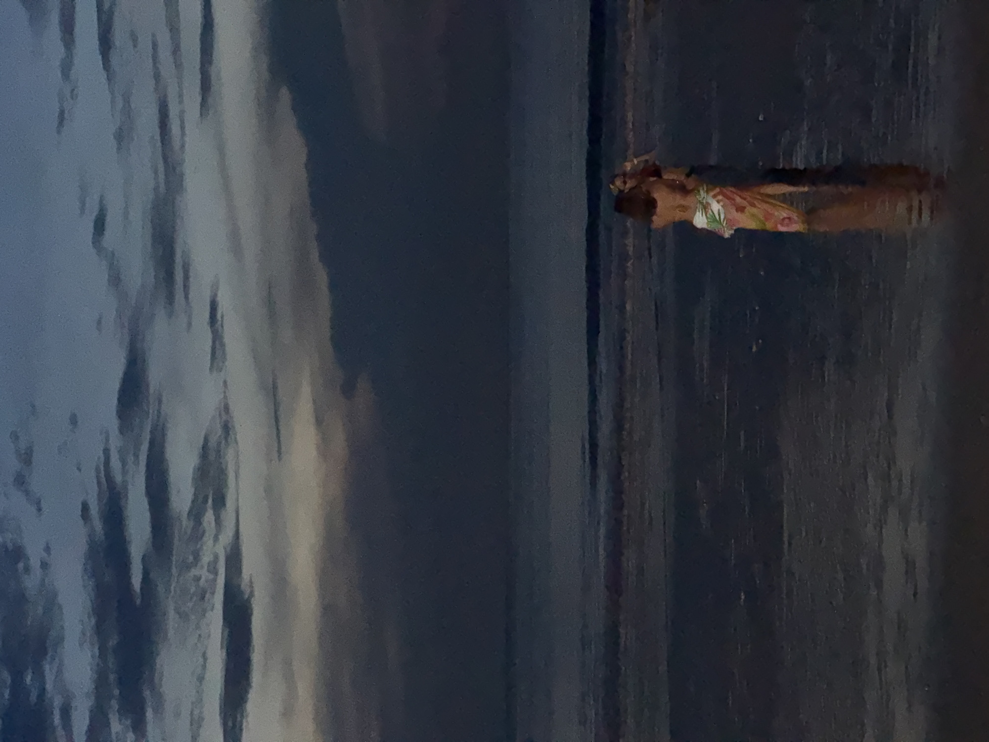 A vertical shot shows a female figure standing in shallow water at an apparent shoreline, possibly in Kuta Selatan, Indonesia, given the prompt. The figure wears a light-colored floral dress. The body reflects on the surface of the water. Behind the figure is a dark expanse of water blending into a cloudy sky.