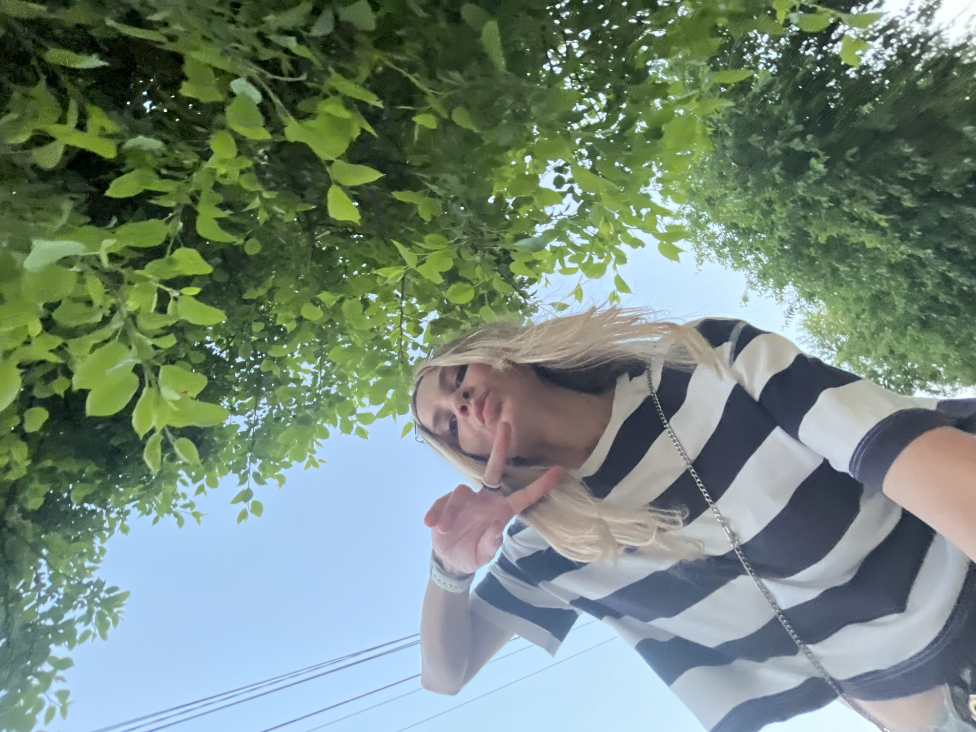 A low-angle, upward-facing photograph shows a blonde woman with a striped shirt posing under trees. She has fair skin and is wearing a silver chain necklace and a wristwatch. She is holding up two fingers in a peace sign. The leaves are green and plentiful, filling the upper portion of the frame. A bright blue sky is visible between the foliage and above the horizon line. Power lines are visible above the horizon. The location is inferred to be in the city of Chaikovsky, Russia.