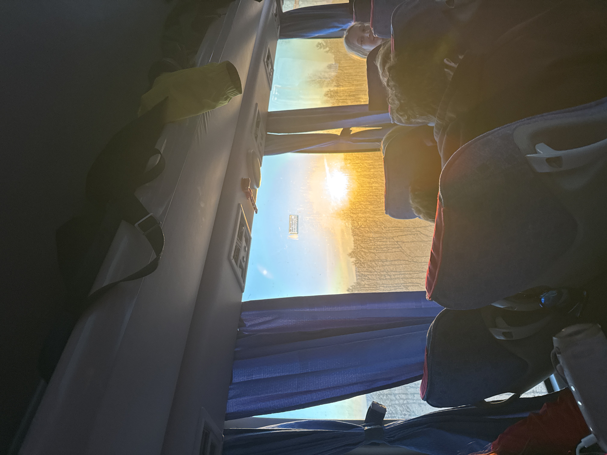 The interior of a passenger bus, possibly a tour bus, is depicted. The bus is partially filled with passengers in various seats. Through the window, bright sunlight is visible along the horizon, with a suggestion of landscape. A curtain partially obscures the view from the window. A yellow backpack and black straps hang from the wall of the bus. A safety notice is affixed on the window. The exact location in the city of Osa, Russia is not discernible from this image.